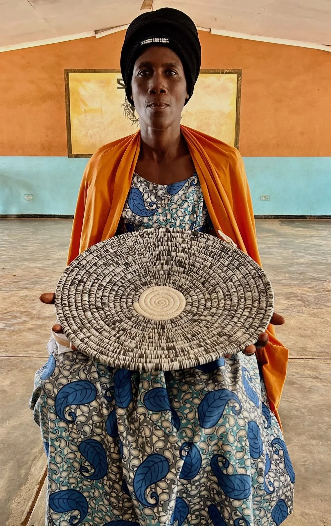A woman in traditional African clothing and a headwrap holding a round woven basket inside a hall with painted walls.
