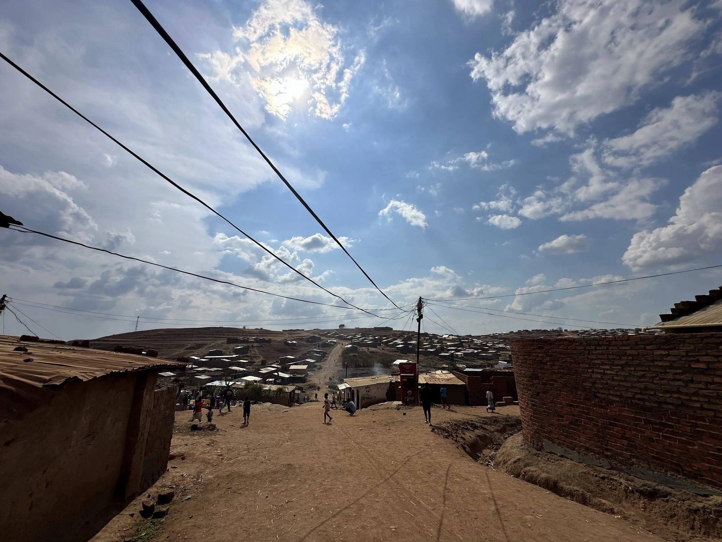 A dirt road in a hilly area with scattered houses and small shops, some buildings with metal roofs, under a partly cloudy sky with sunlight and electrical wires running overhead.
