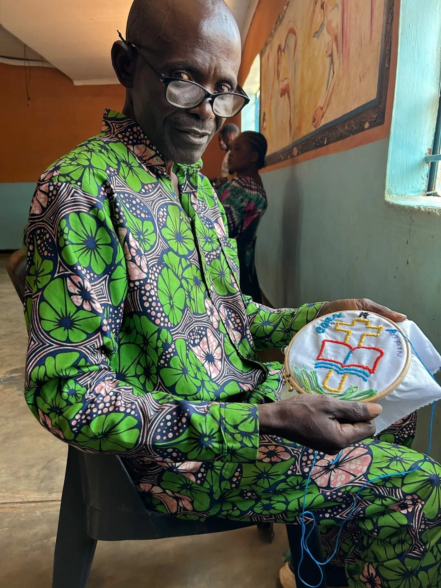 A man wearing glasses and a colorful green and pink patterned shirt is sitting and holding an embroidery hoop with a design that includes a cross, a book, and the word 'God' in a church or community center. Two women are visible in the background near a wall with a painting or mural.