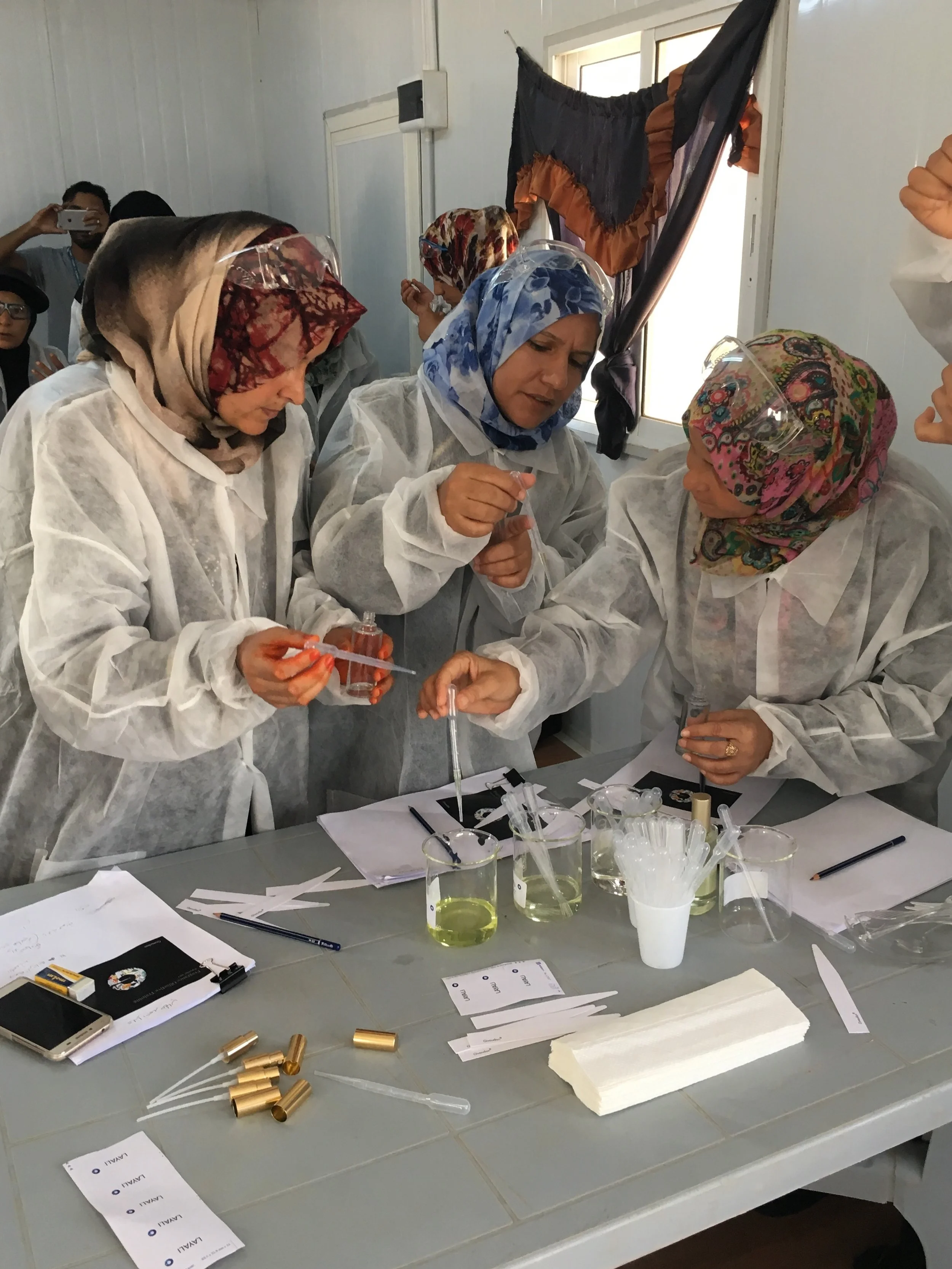 Women in protective lab coats and colorful headscarves conducting a chemistry experiment with test tubes, liquids, and laboratory equipment on a table.
