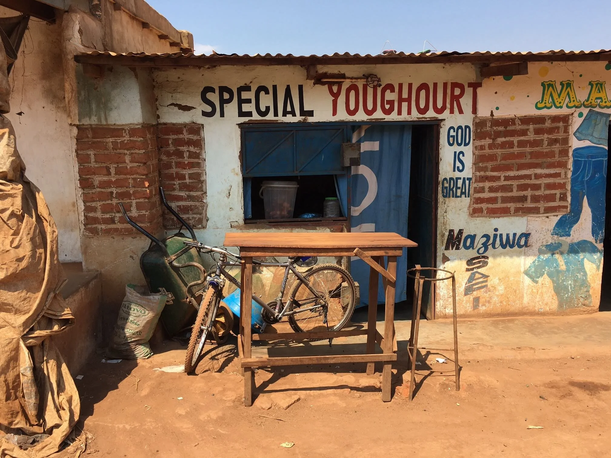 Small roadside shop with a sign that reads 'SPECIAL YOGHOURT' and 'GOD IS GREAT'. In front of the shop, a wooden table, a bicycle, and a metal stand are visible. The shop has partially exposed bricks and a blue curtain covering the entrance.