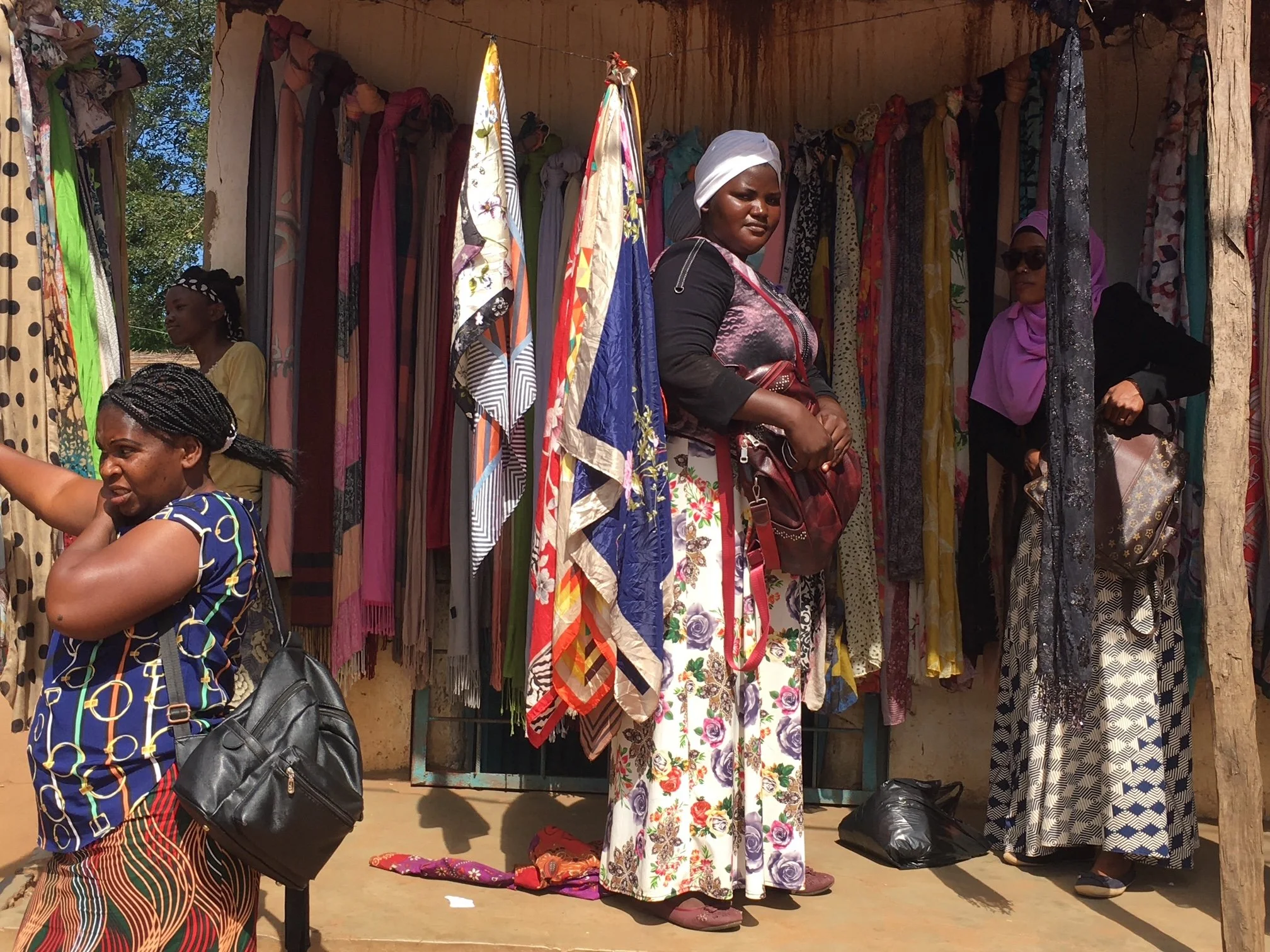 Women shopping at an outdoor stall with colorful scarves and clothing hanging.