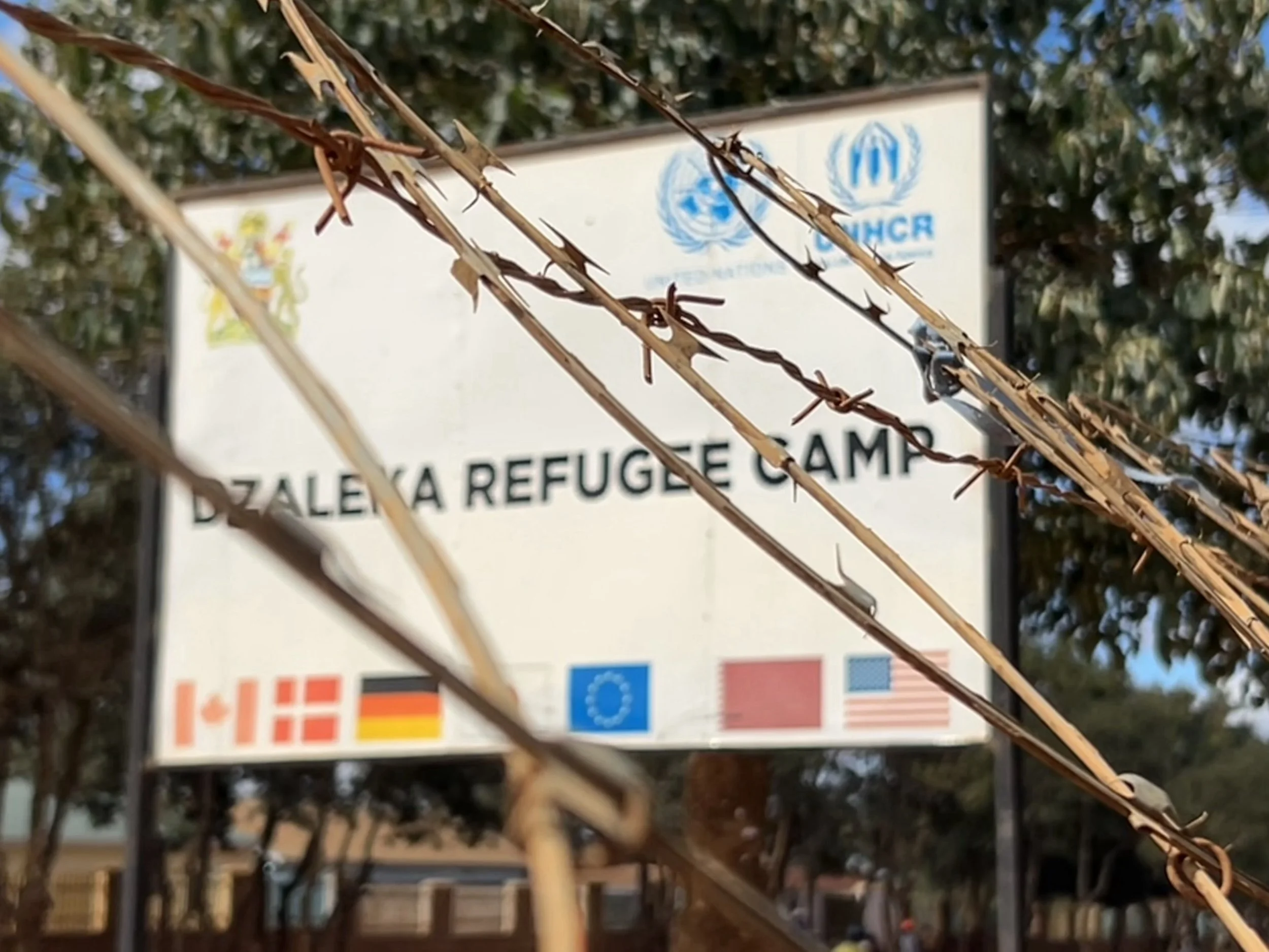 Barbed wire in the foreground with a blurred sign reading 'Dzaleika Refugee Camp' and the UNHCR logo in the background.
