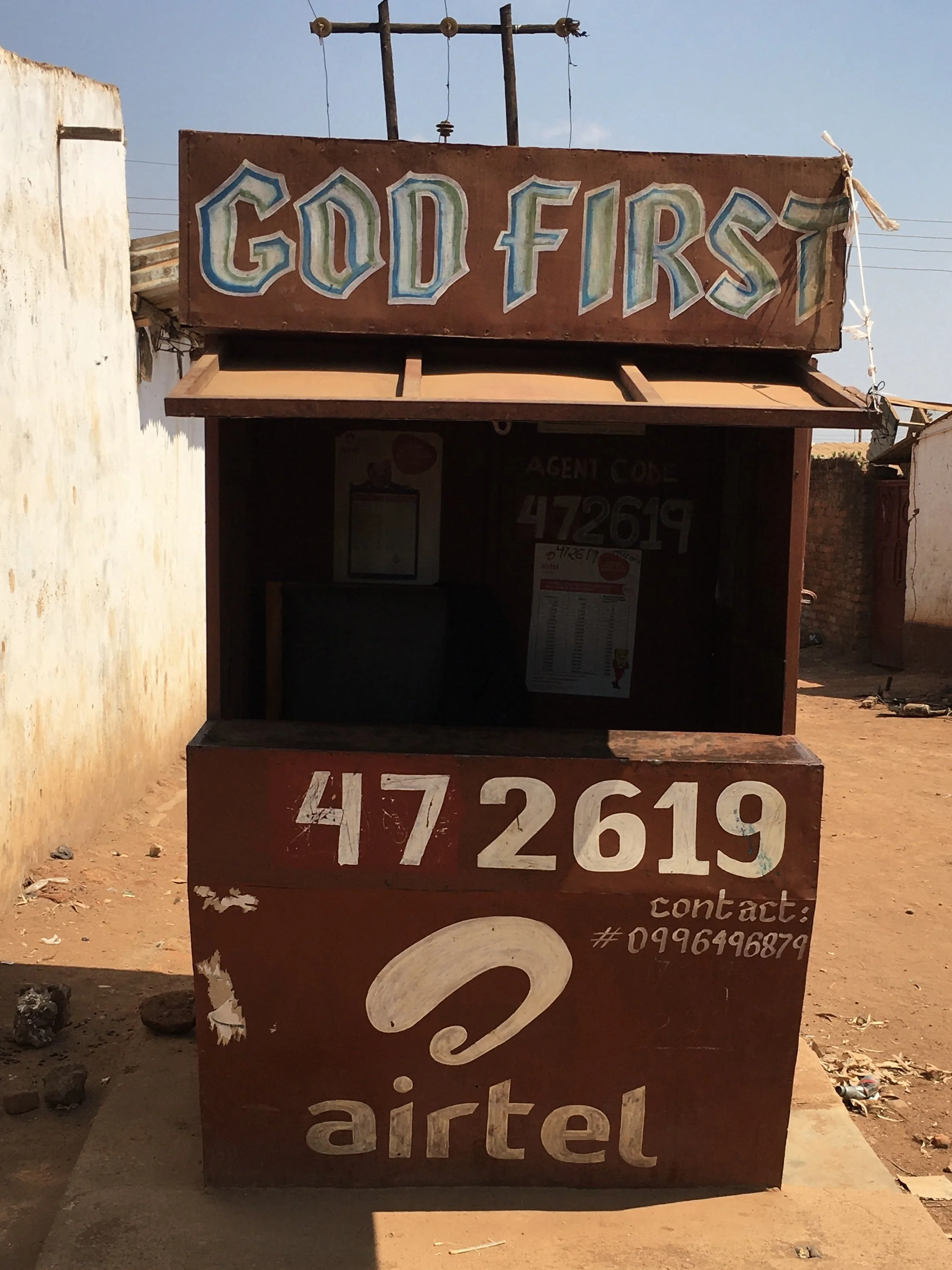 A small roadside kiosk with a sign that reads 'GOD FIRST' on top. The kiosk has a contact number and the Airtel logo on the front.