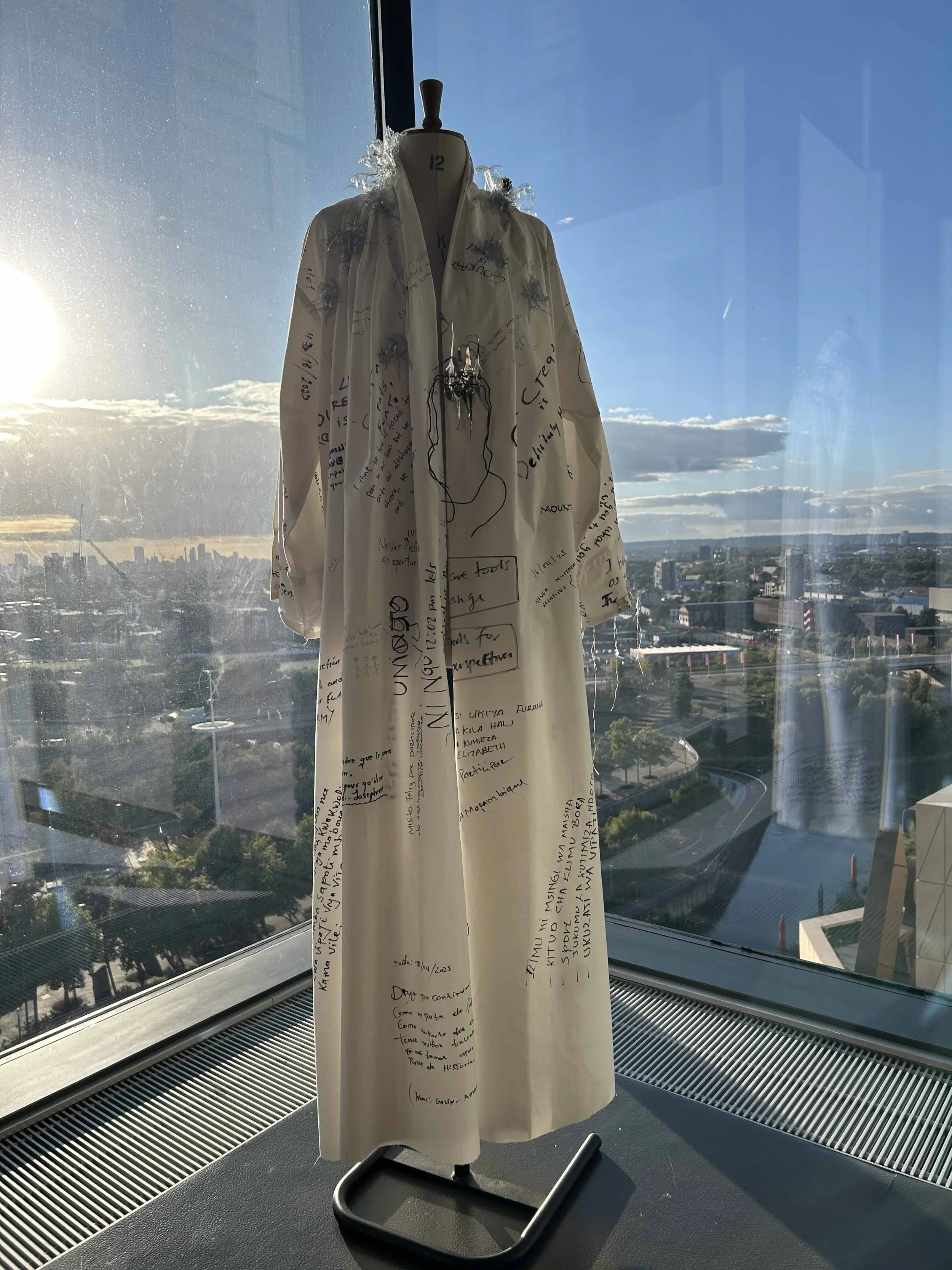 A mannequin dressed in a long white coat with handwritten notes and sketches all over it, displayed indoors near a large window with a cityscape and blue sky in the background.