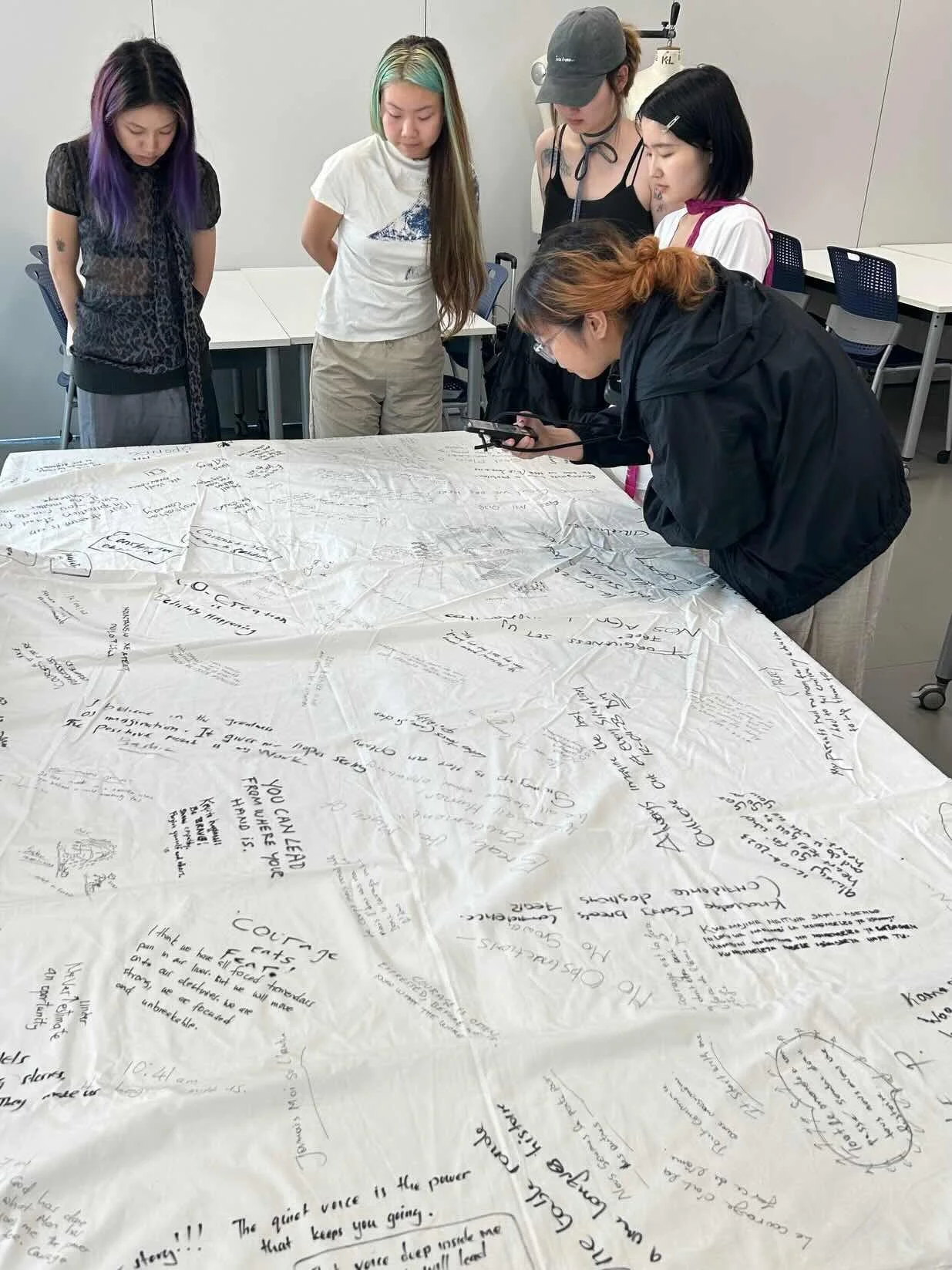 Five women gather around a large white cloth on a table, which is covered in handwritten notes and drawings. One woman is writing with a marker, while the others watch.