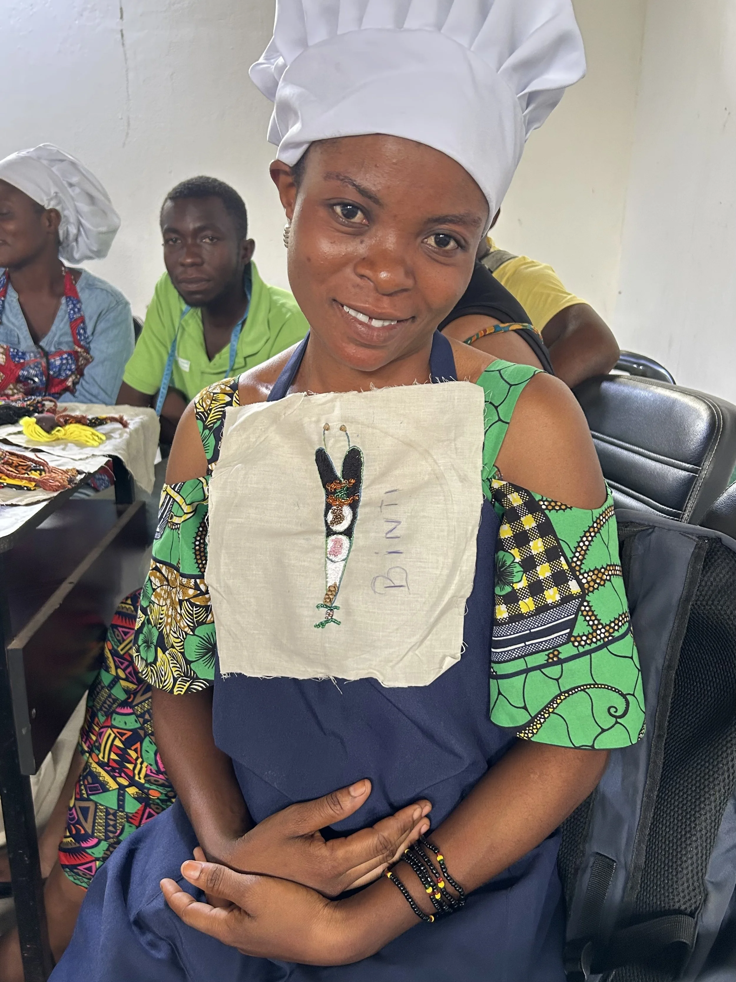 A woman wearing a white chef's hat and colorful traditional clothing holds a piece of fabric with a hand-stitched beaded bracelet design and the name 'BINI' embroidered on it.