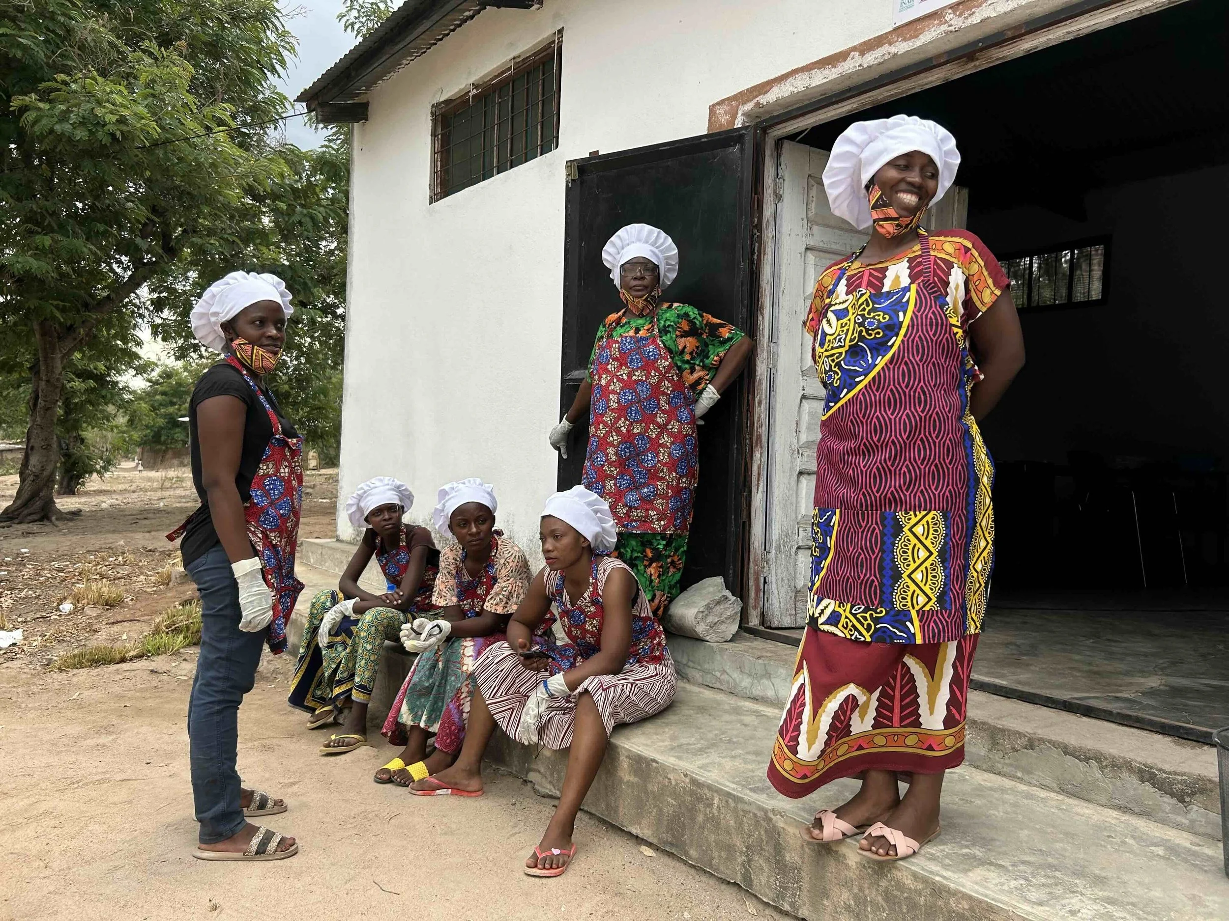 Group of women and girls gathered outside a building, wearing colorful traditional African attire and headscarves, some sitting on the ground and others standing, smiling and posing for the photo.