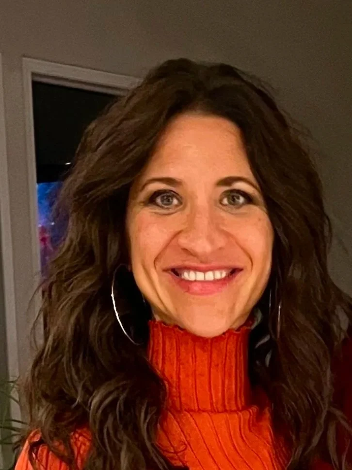 A woman with long curly brown hair smiling, wearing a red-orange sweater, in an indoor setting with a doorway behind her.