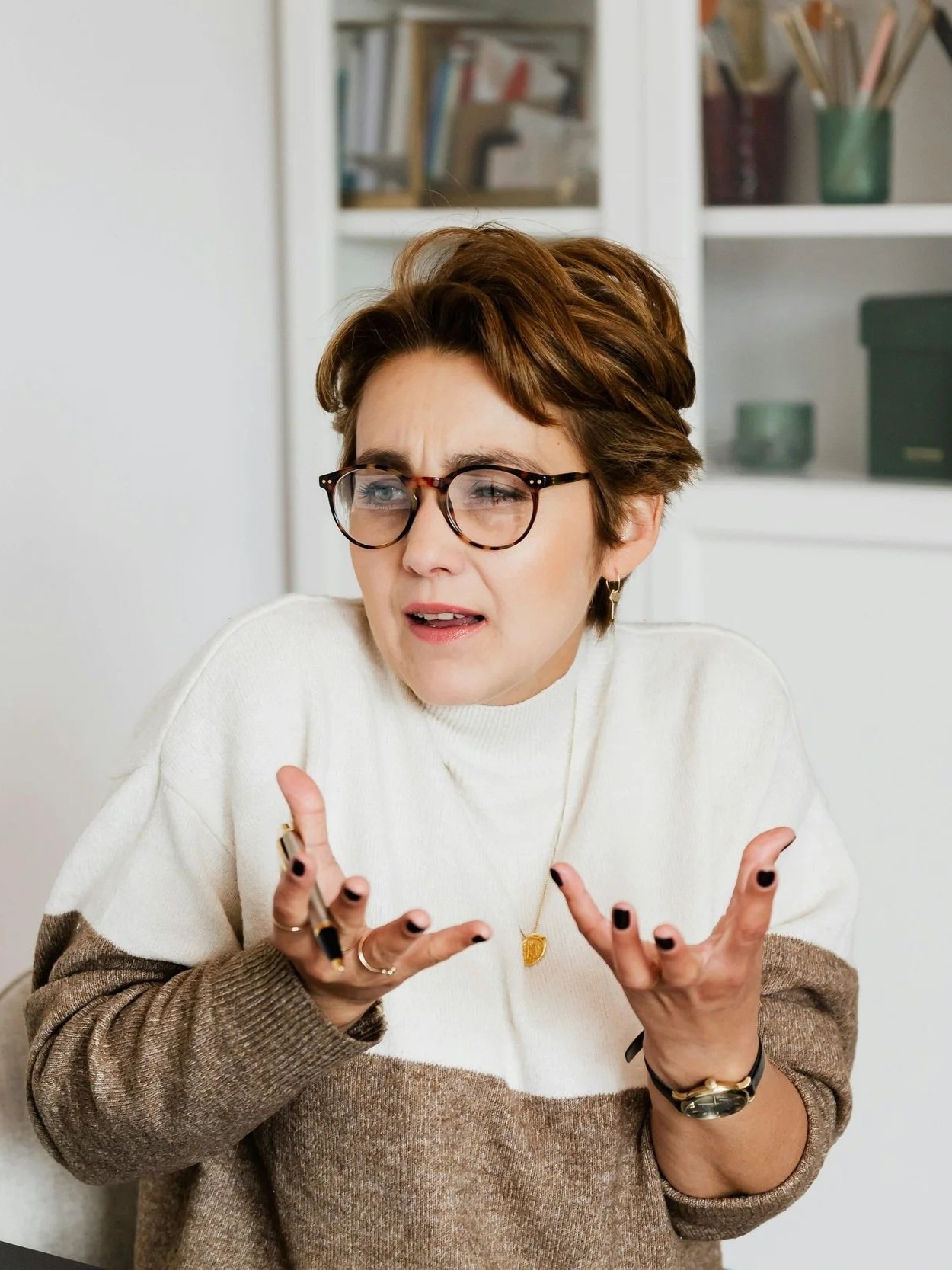 A woman with short brown hair and glasses, wearing a white and brown sweater, appears confused or surprised while holding a pen and gesturing with her hands in front of a bookshelf.