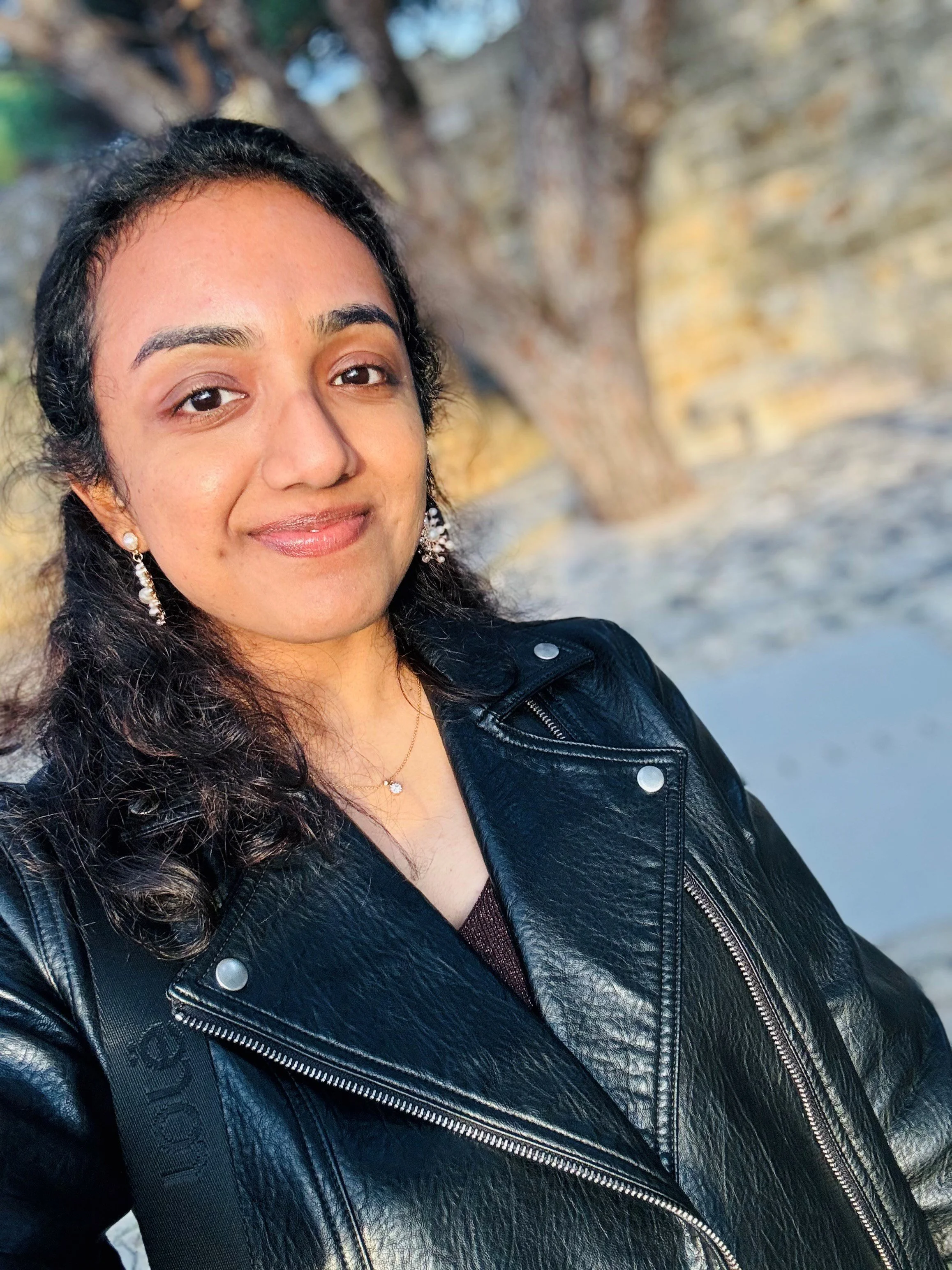 A woman smiling outdoors with trees in the background, wearing a black leather jacket, earrings, and a necklace.