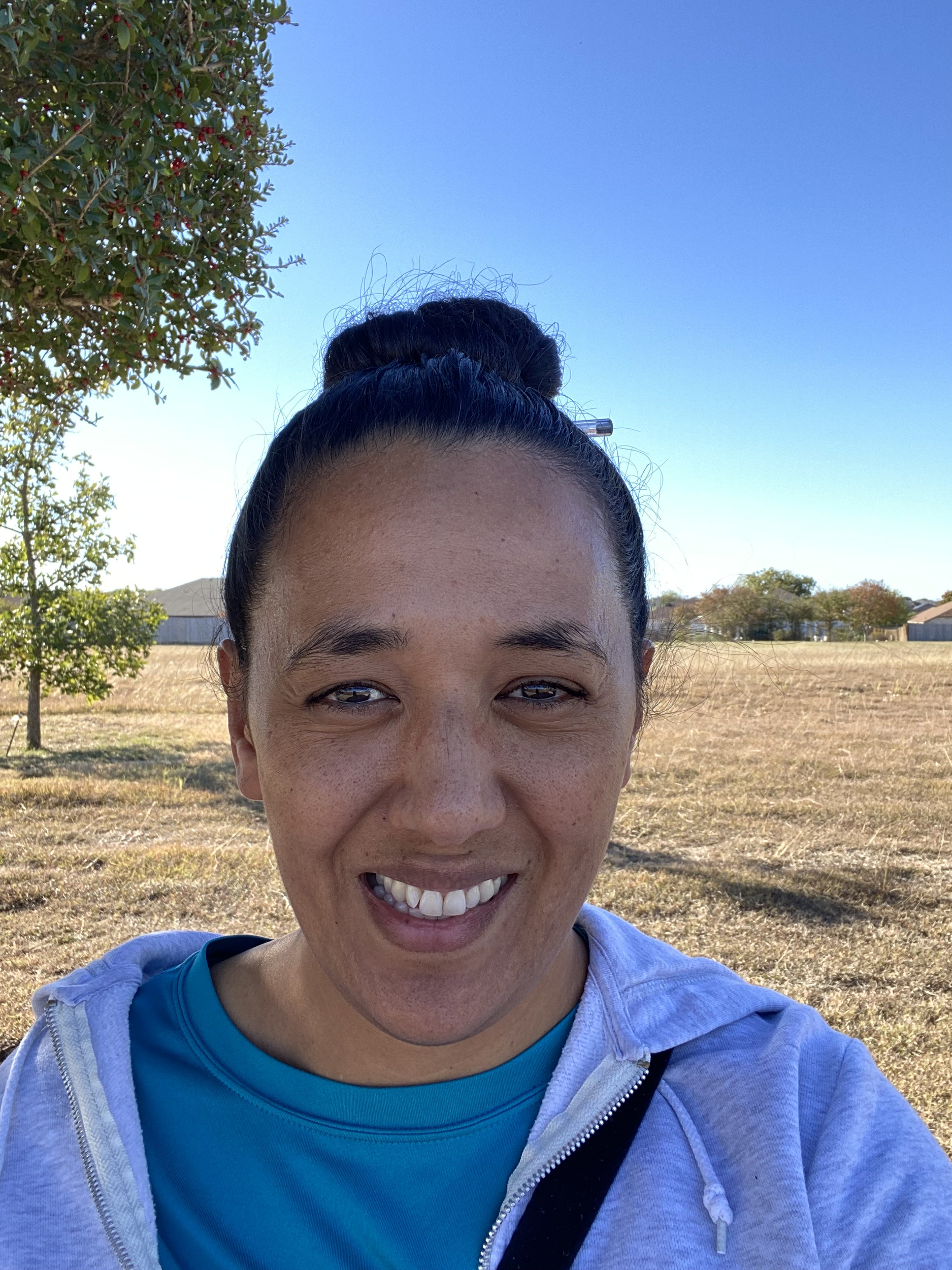A woman smiling outdoors in a park on a clear, sunny day. She has dark hair tied in a bun, freckles, and is wearing a blue shirt and light gray hoodie.