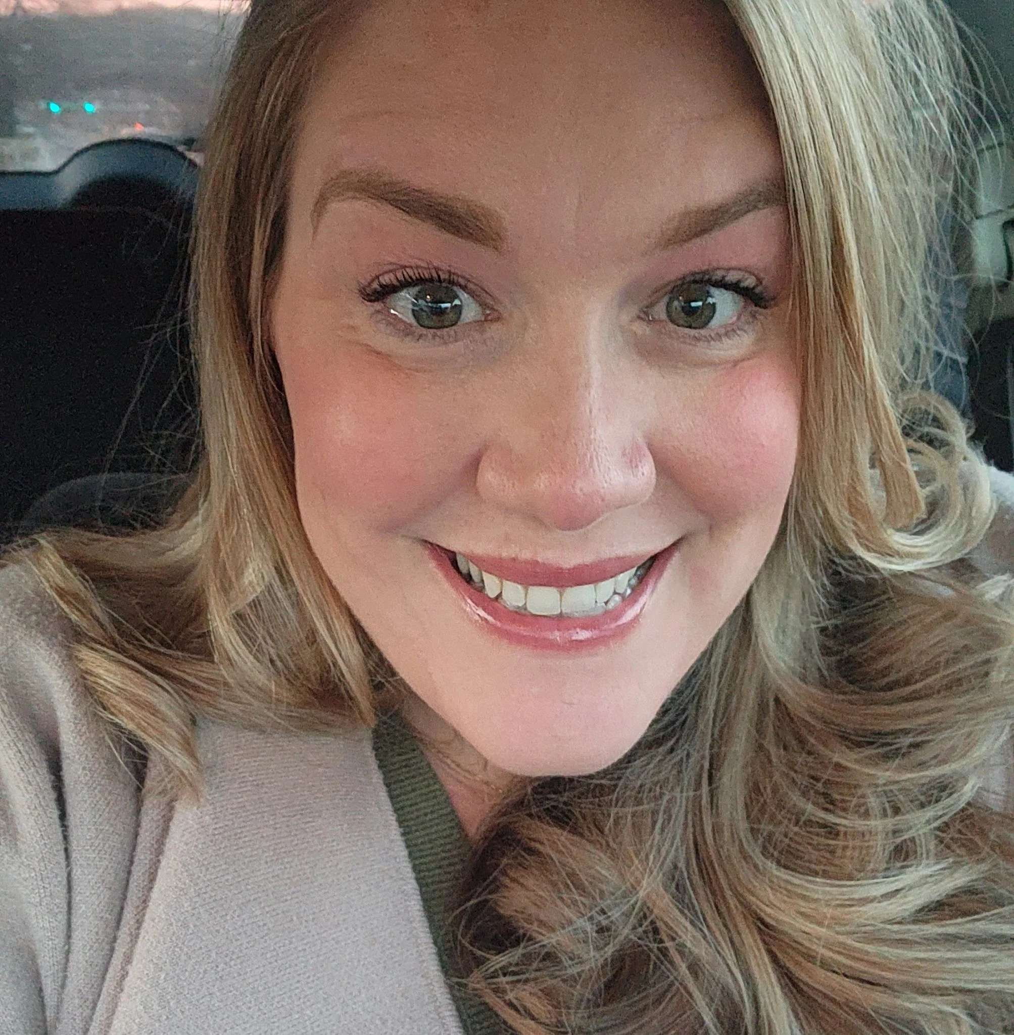 Close-up of a smiling woman with long wavy blonde hair, sitting in a car, with blurred background showing other vehicles.