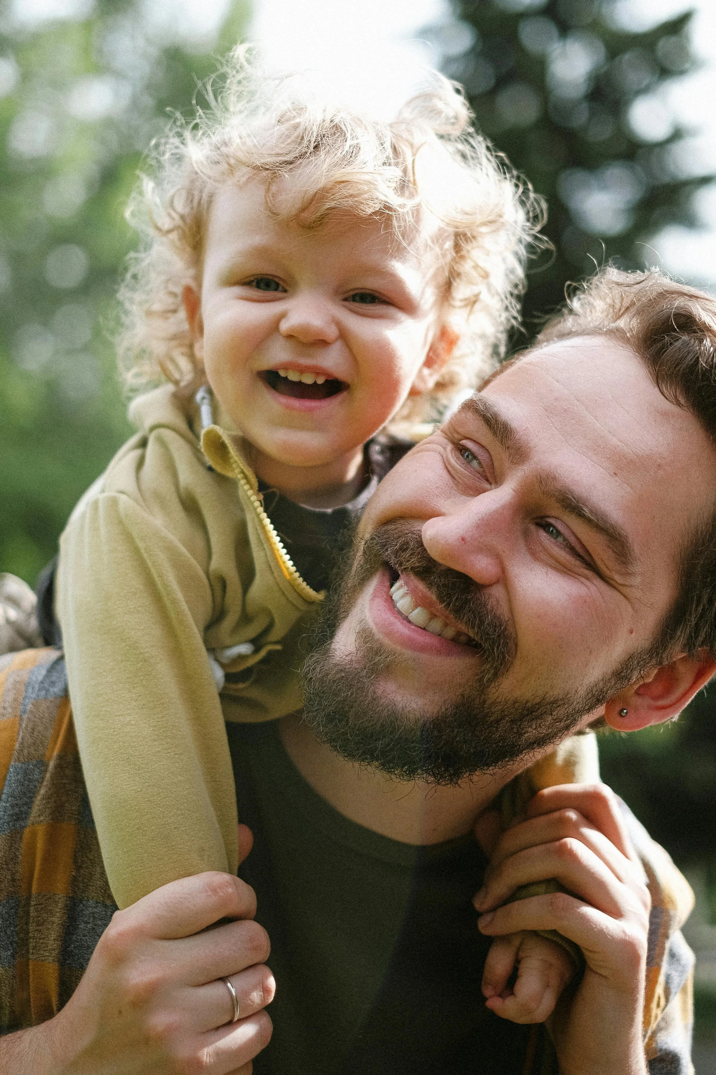 A young child with curly blonde hair smiling while riding on an adult man's shoulders outdoors surrounded by trees.