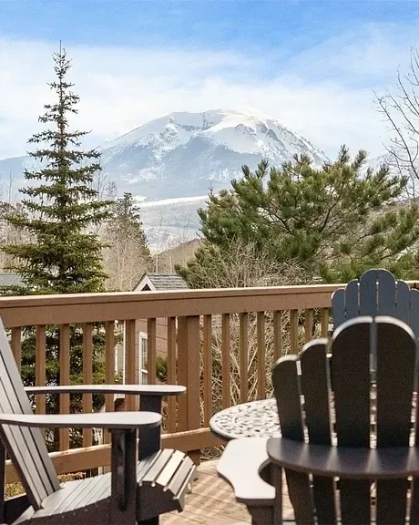 View of a snow-capped mountain in the distance, seen from a wooden deck with outdoor furniture, surrounded by trees.