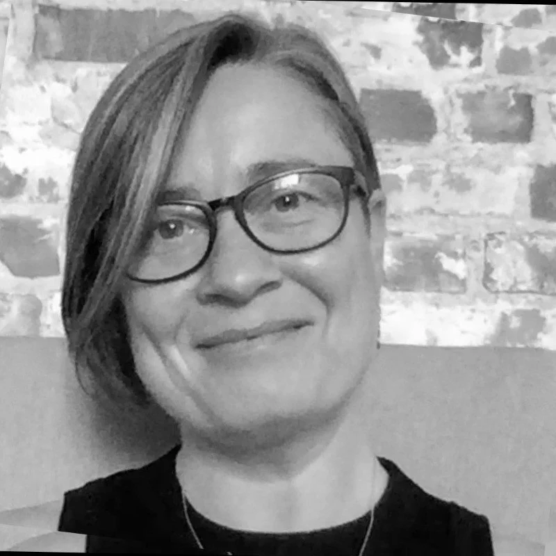 A woman with short hair and glasses smiling, sitting against a brick wall background.