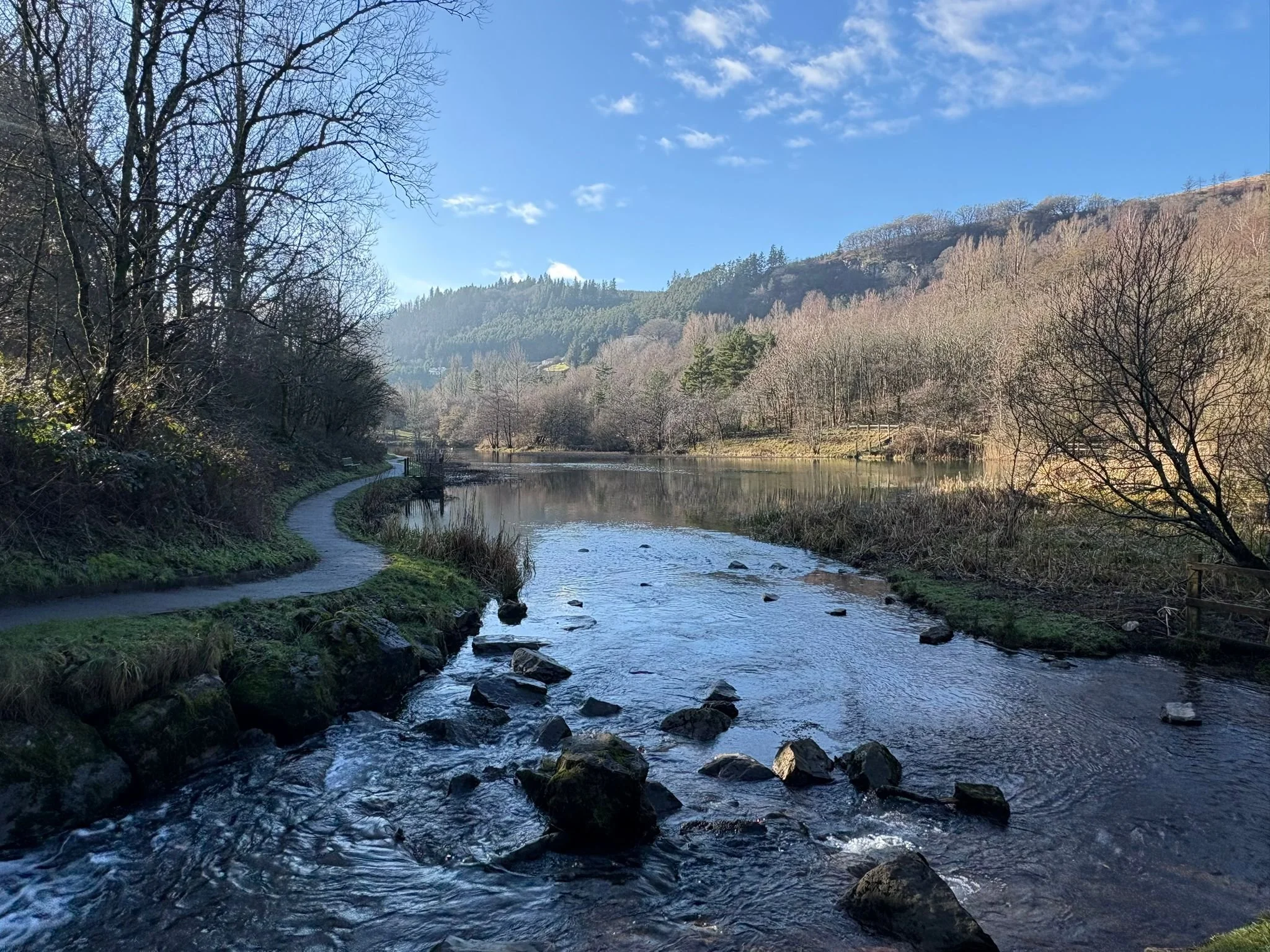 A peaceful river scene with a winding pathway on the left side, bare trees along the riverbank, and hillside with trees in the background under a blue sky with some clouds.
