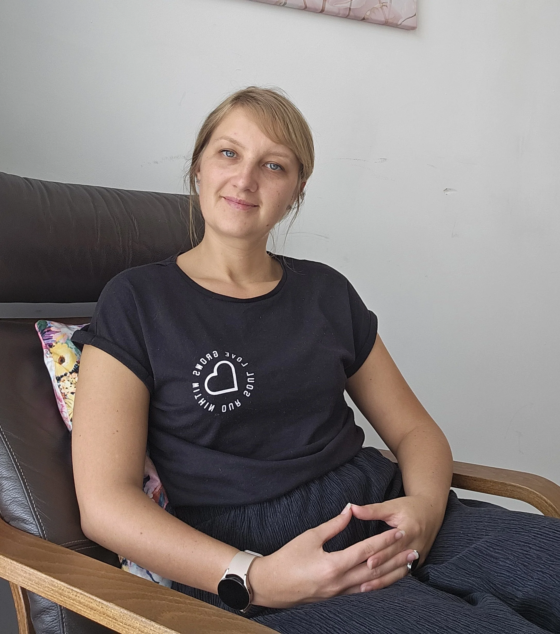 A woman with blonde hair and blue eyes sitting on a wooden chair with a dark cushion, wearing a black t-shirt and dark pants, smiling softly.