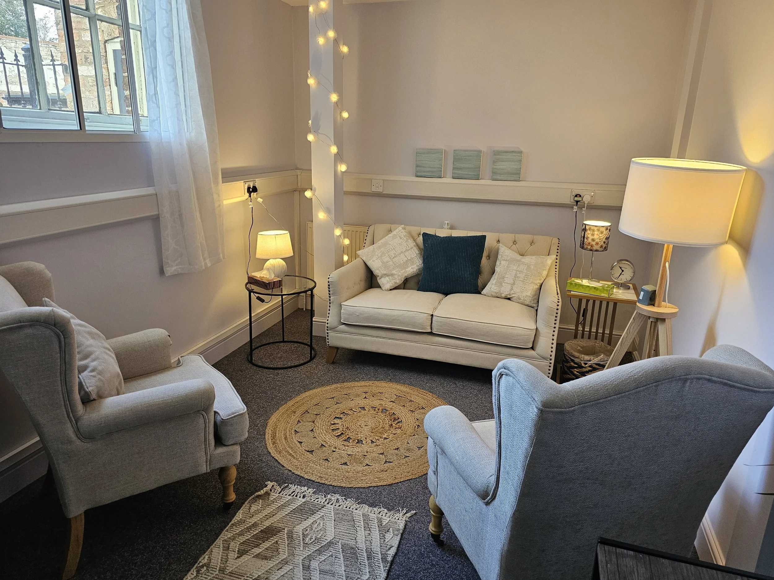 A cozy living room with a white sofa adorned with pillows, two armchairs, a round woven rug, two side tables with lamps, a string of fairy lights, and a window with sheer curtains.