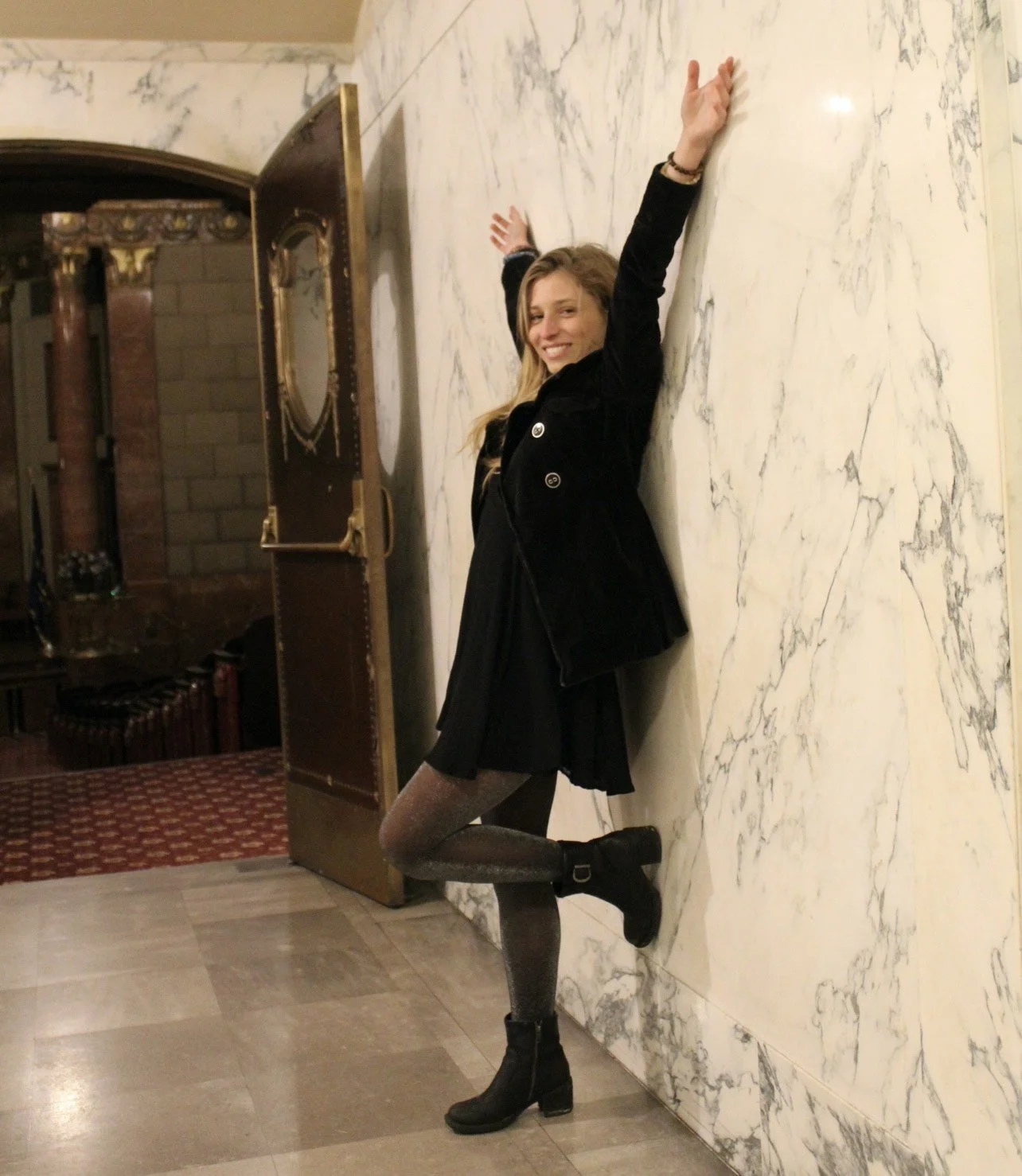 A woman with long blonde hair smiling and stretching her arms up, leaning against a white marble wall inside a building with luxurious decor.