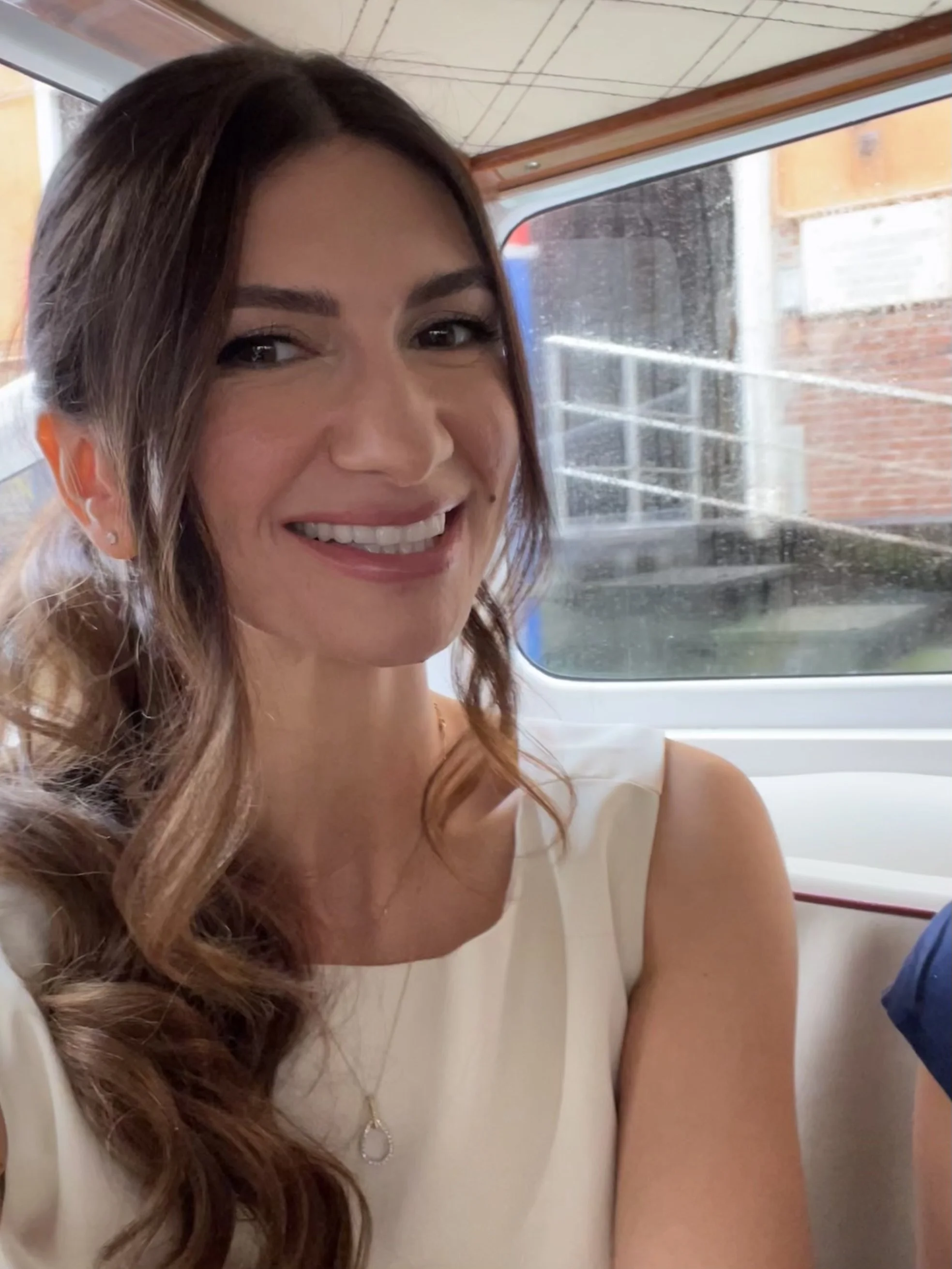 A woman with long wavy brown hair and a white top, smiling while sitting inside a vehicle with a window behind her showing rain outside.