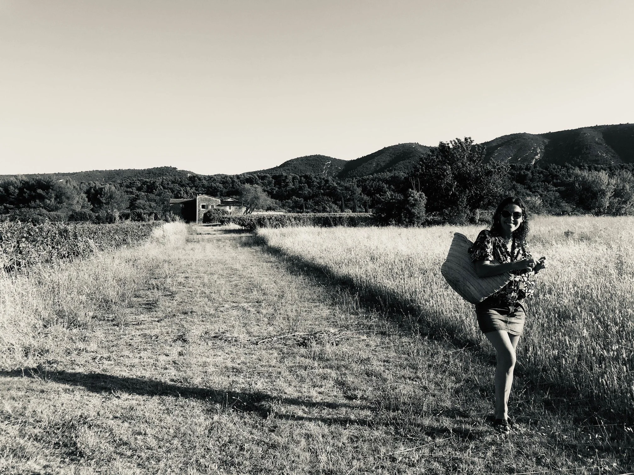 Une femme portant des lunettes de soleil, un sac de plage, une robe à motifs et des sandales marche sur un chemin rural dans un champ. En arrière-plan, il y a des montagnes et des arbres.