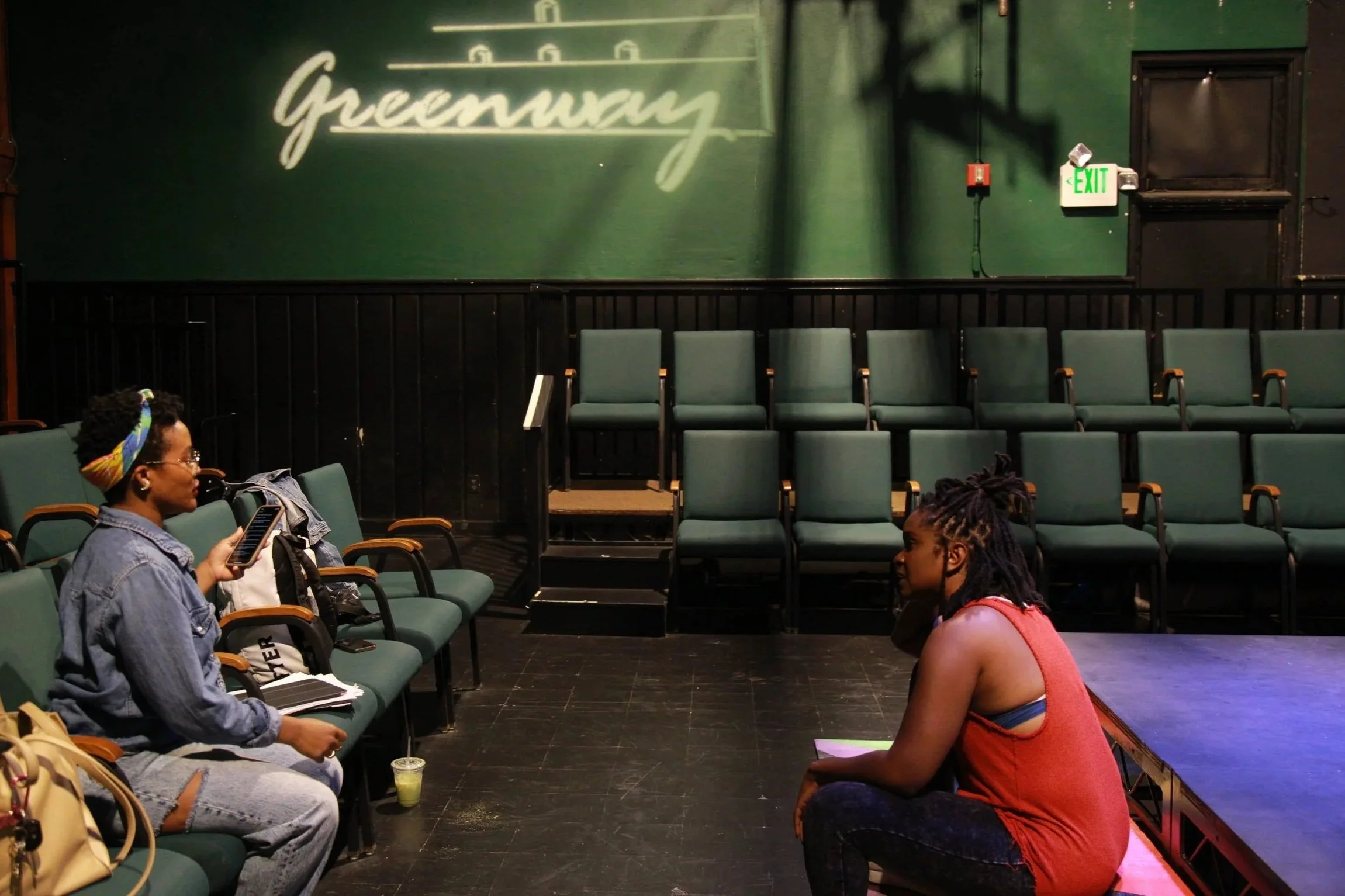 Two women are sitting and facing each other in a theater with rows of empty green seats behind them. One woman is on the left, using her phone, wearing glasses, a denim jacket, and a headscarf, with a drink and a bag nearby. The other woman is on the right, sitting on the stage, wearing a red sleeveless top and dark pants, with her hair in dreadlocks. There is a green wall in the background with a cursive sign that reads "Greenway" and an emergency exit sign.