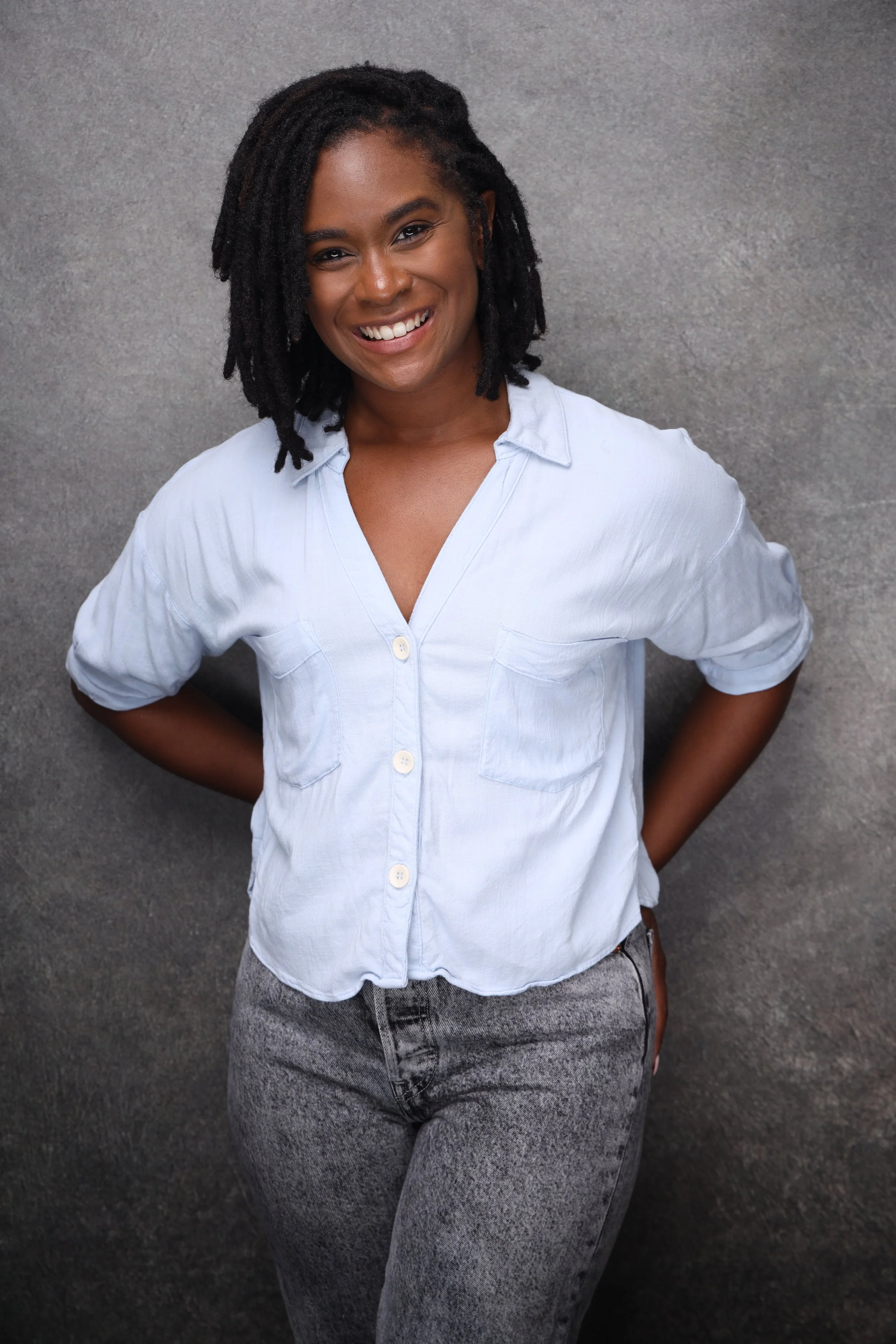 A young woman with locs smiling, wearing a white button-up shirt and gray acid-washed jeans, standing against a gray textured background.