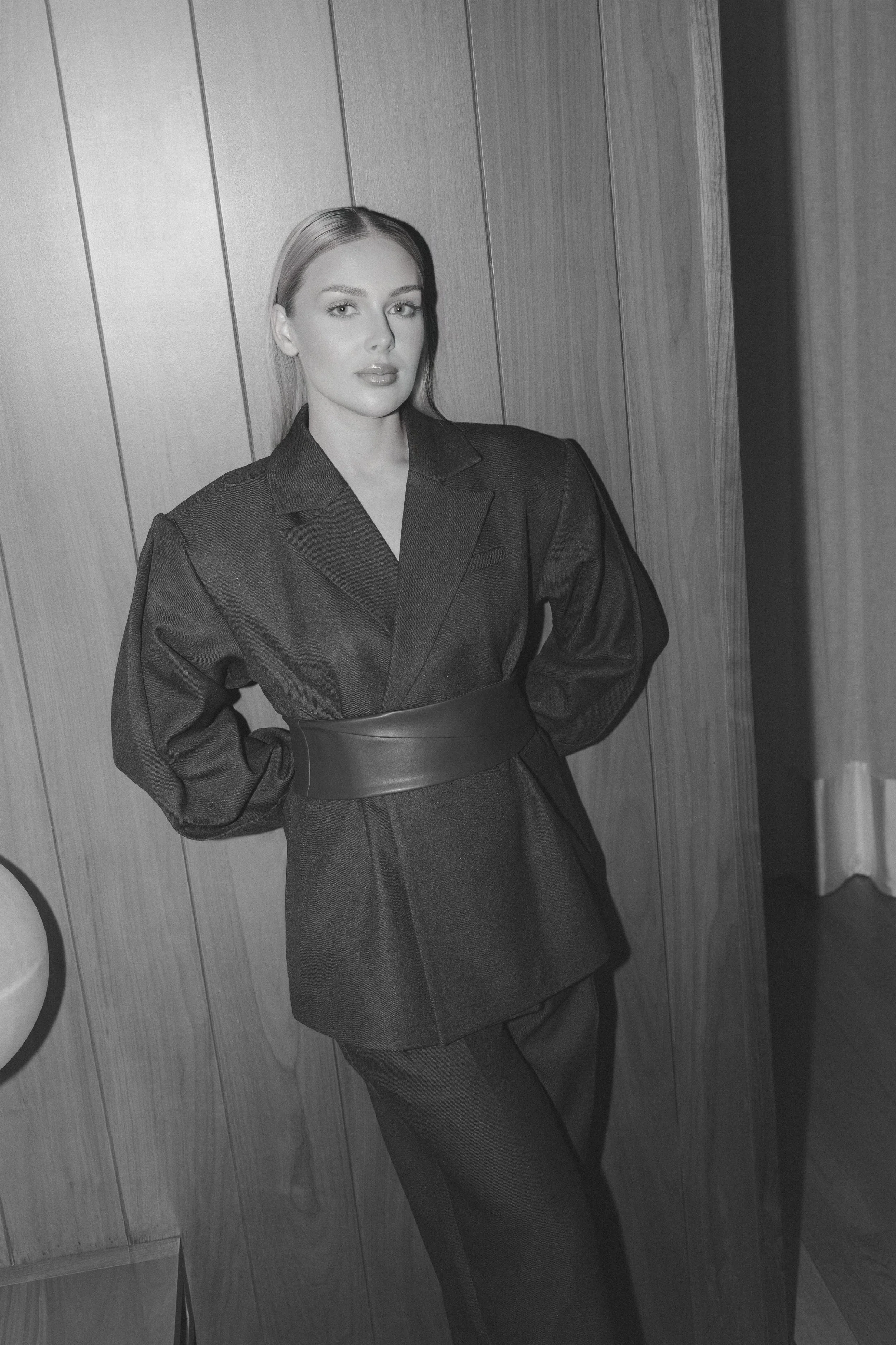 Black and white photograph of a woman with long hair, wearing a dark blazer and matching pants, standing against a wooden paneled wall.