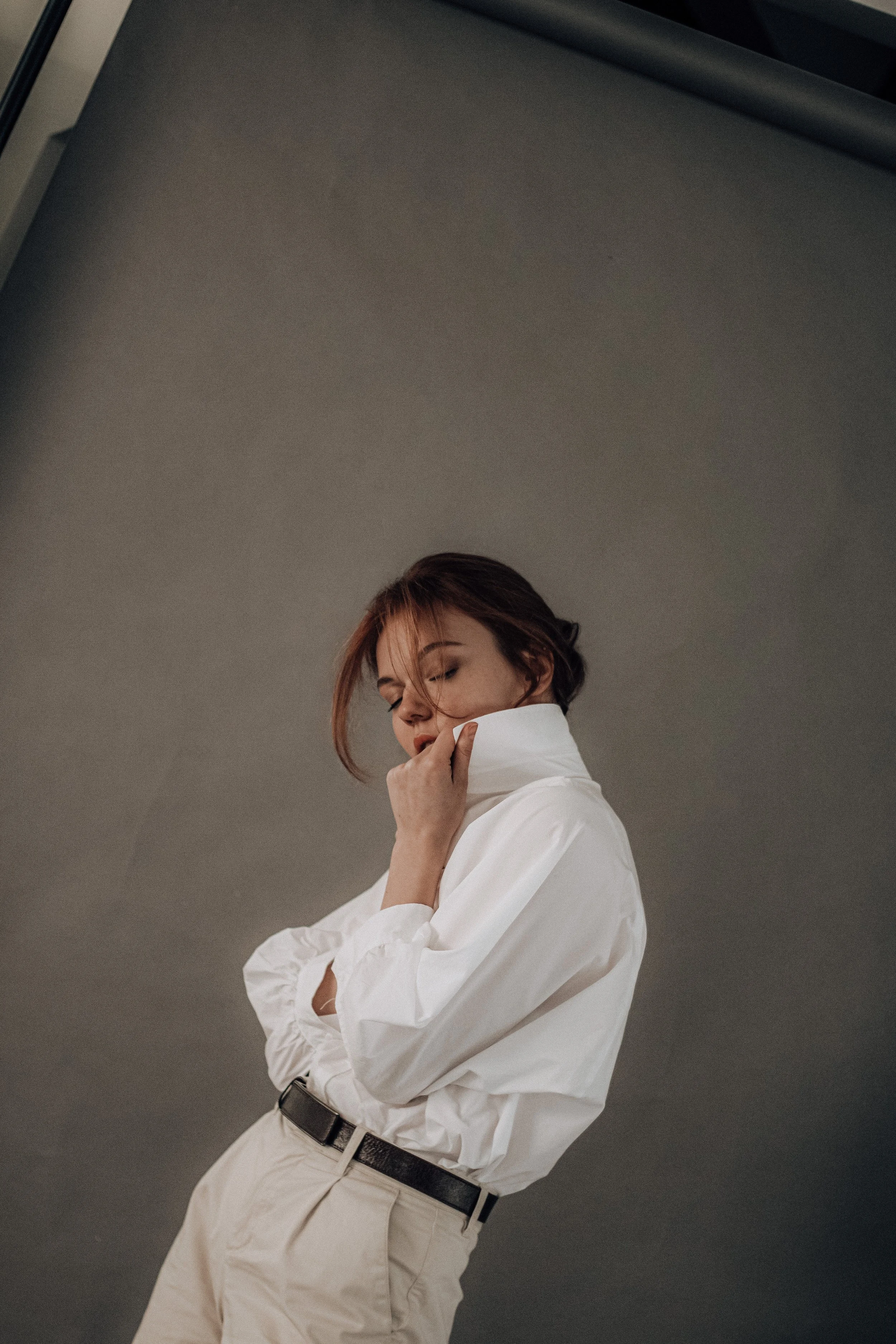 A woman with brown hair wearing a white shirt and beige pants, standing against a gray wall, with her eyes closed and hand touching her face.