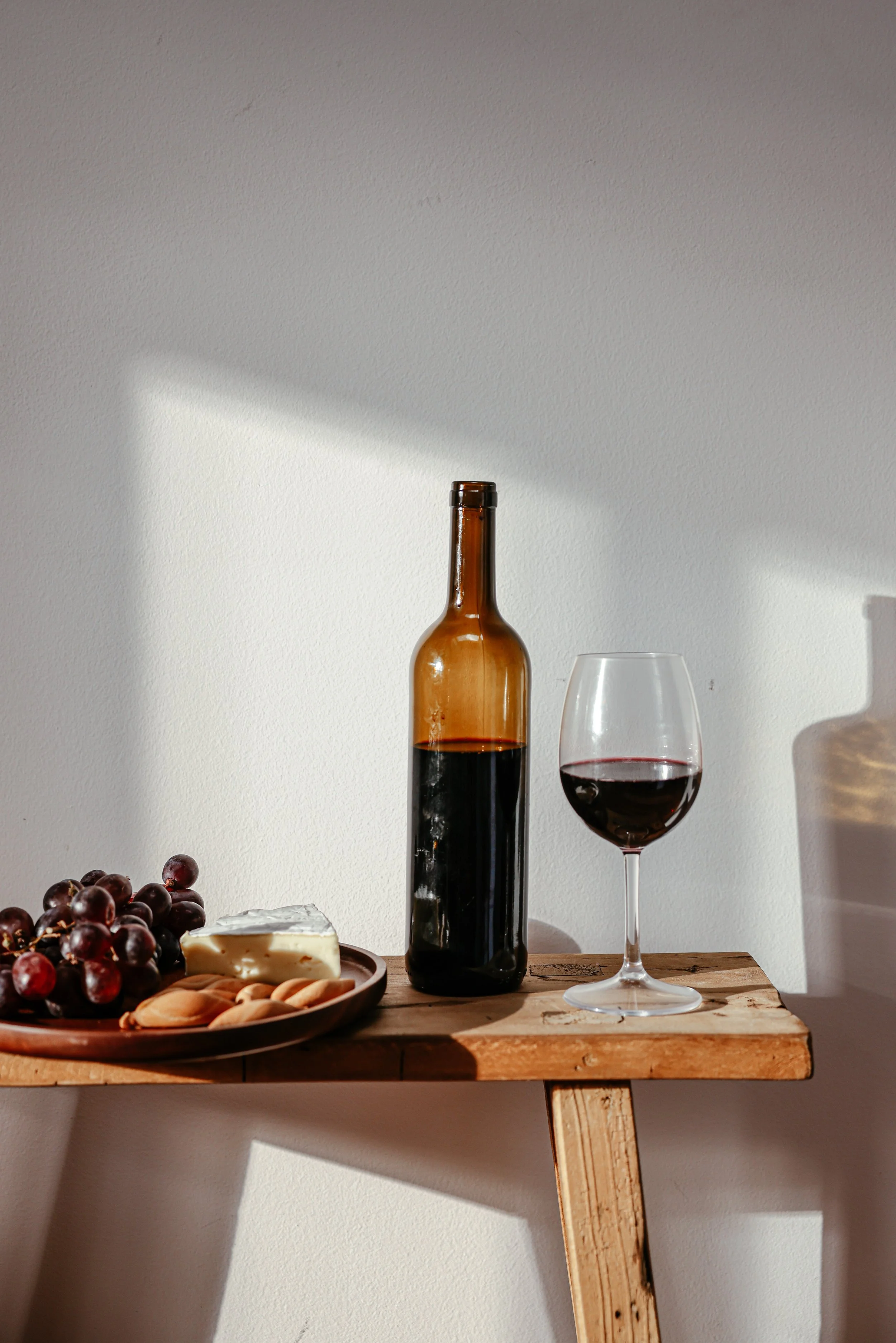A wooden table holds a wine bottle, a glass of red wine, a plate with grapes, cheese, and crackers, with sunlight casting shadows on a white wall.