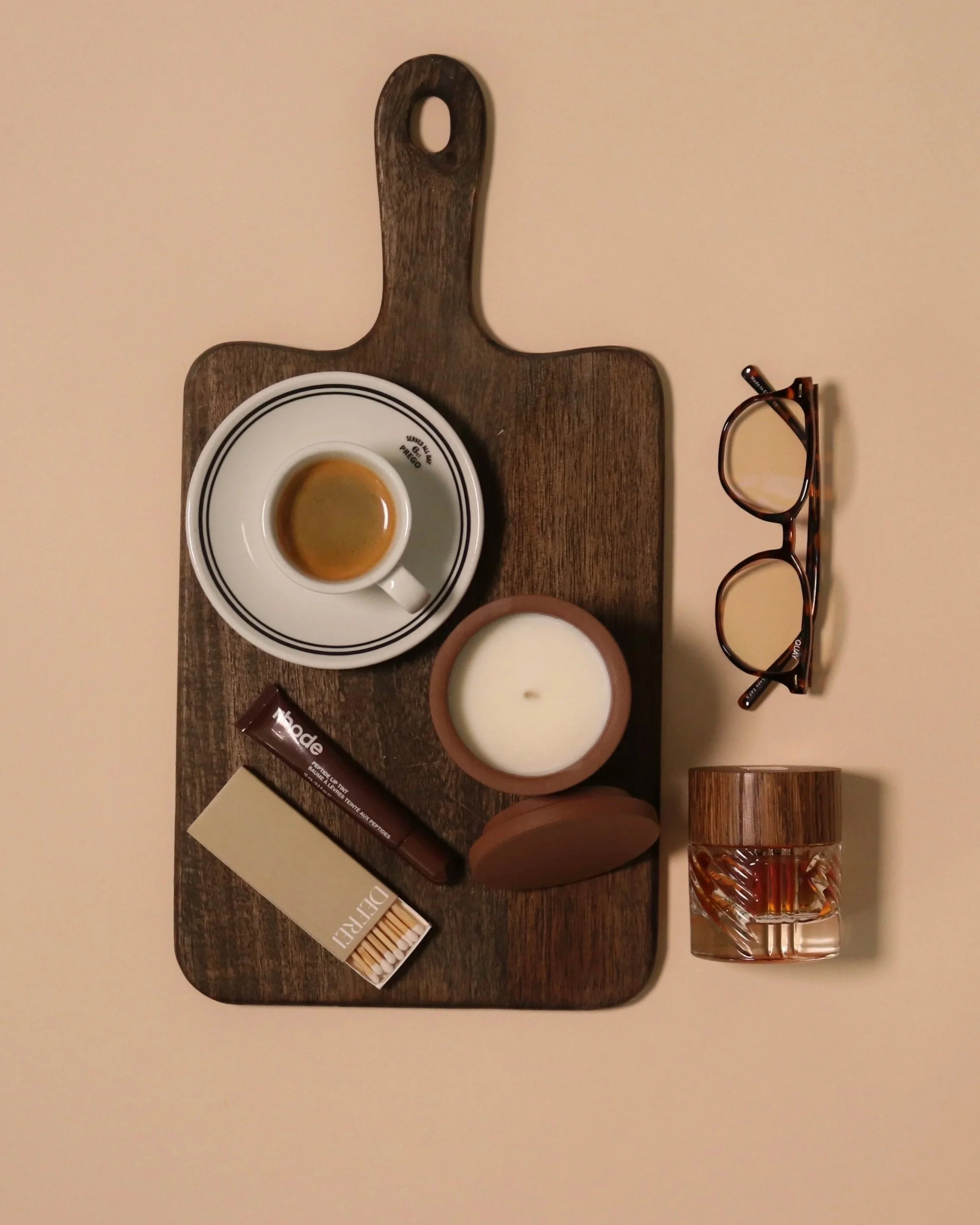 A wooden serving board with a shot of espresso, a lit candle in a terracotta holder, a chocolate bar, a box of matches, a glass of water, and a pair of glasses placed on a beige surface.