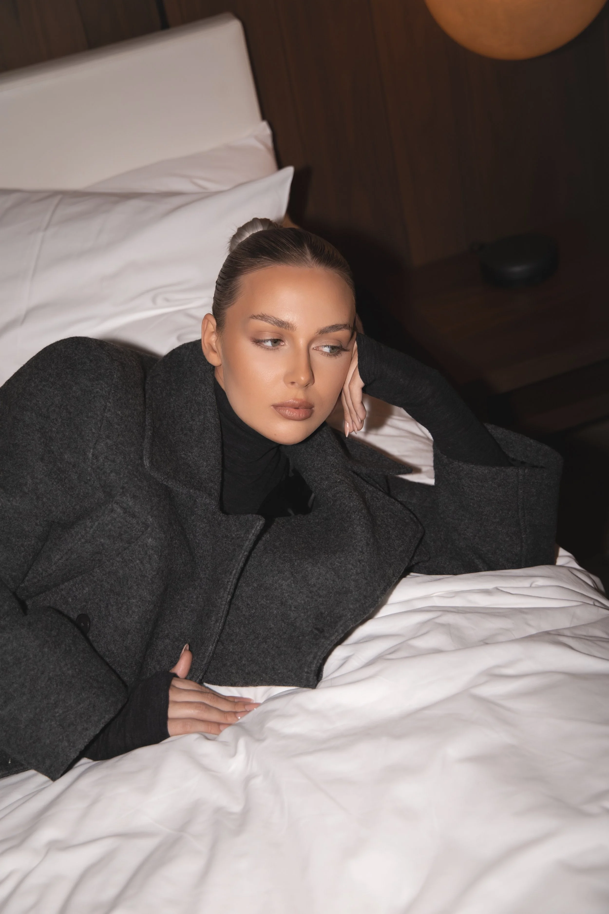 A woman with blonde hair in a bun lying on a bed with white sheets, wearing a dark gray coat and black turtleneck, resting her head on her hand and looking to the side.