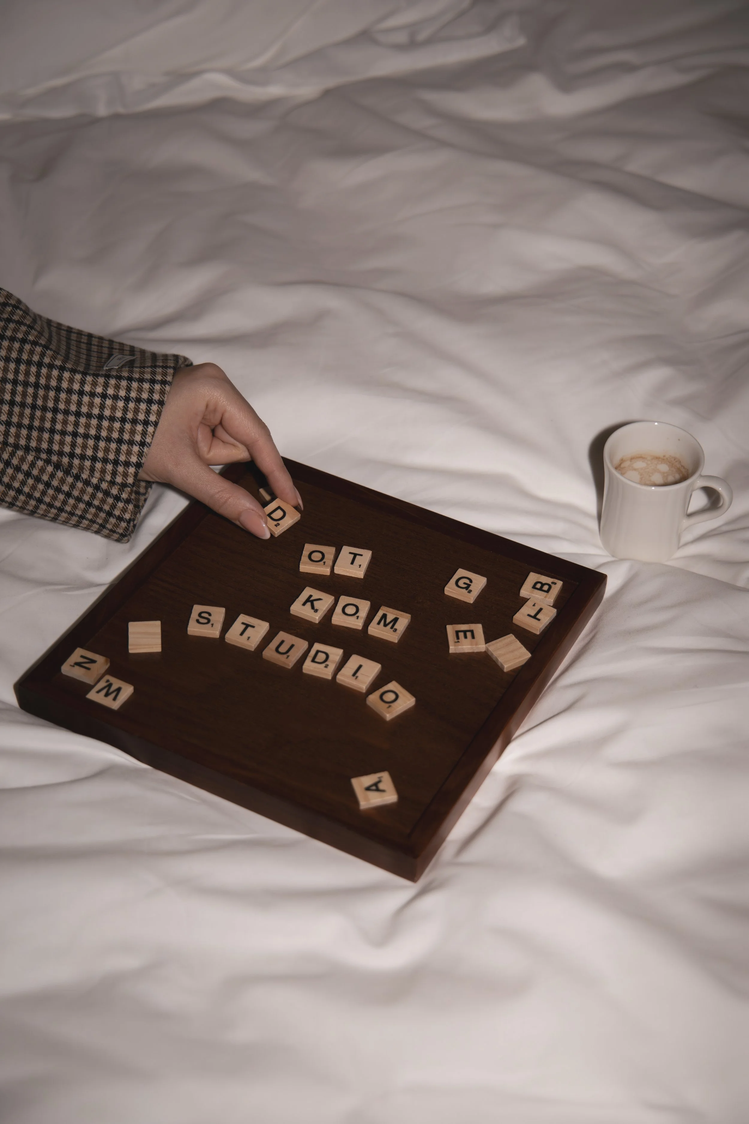 A hand with a checkered shirt is arranging Scrabble tiles on a dark wooden tray. Some tiles spell out words like 'STUDIOS' and 'KOM'. There is a mug of coffee or hot chocolate nearby on a white sheet.