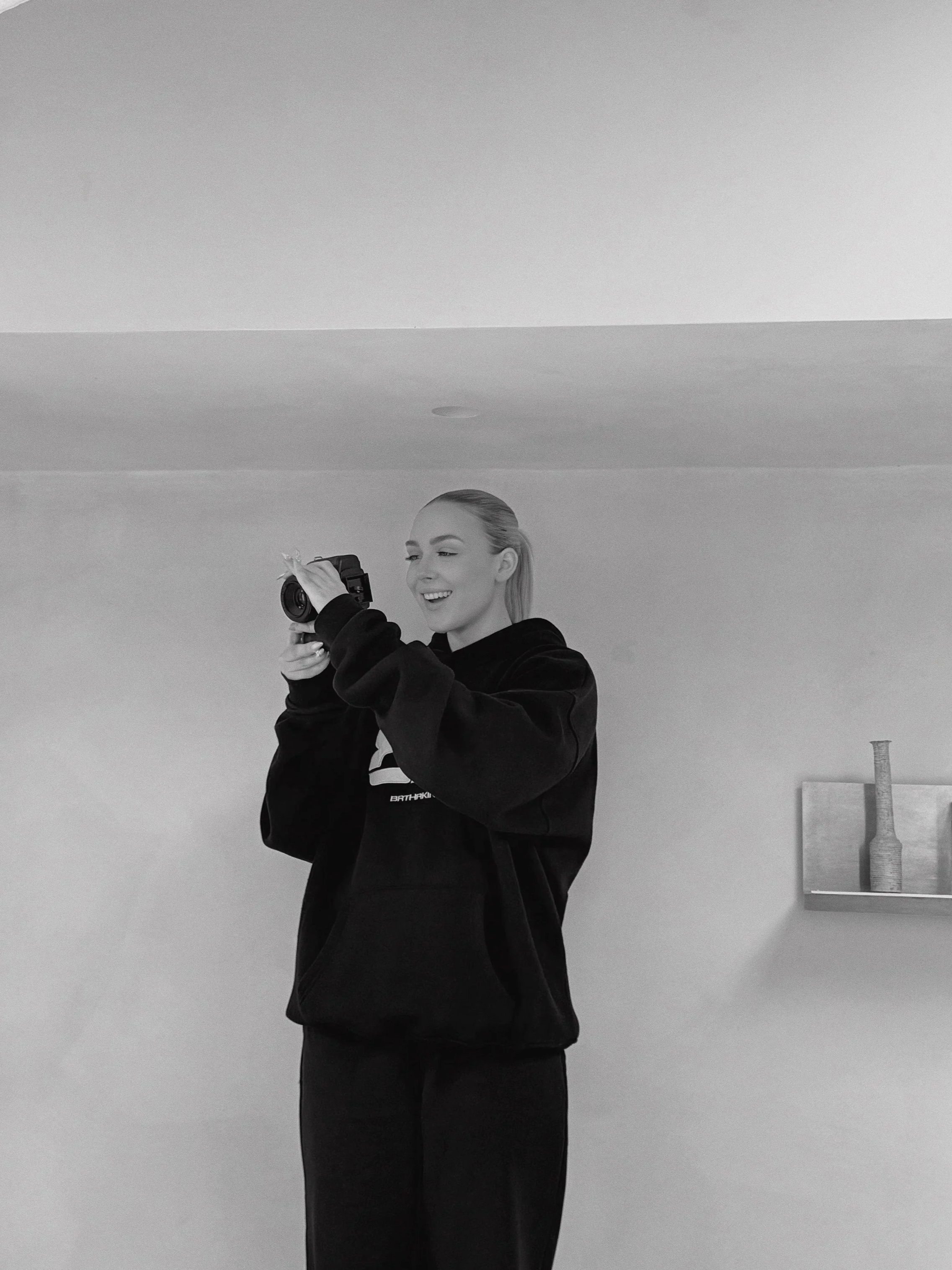 A young woman with blonde hair in a ponytail, wearing a black hoodie and black pants, is taking a photo with a camera in a minimalistic room with a plain wall and a small shelf holding decorative vases.