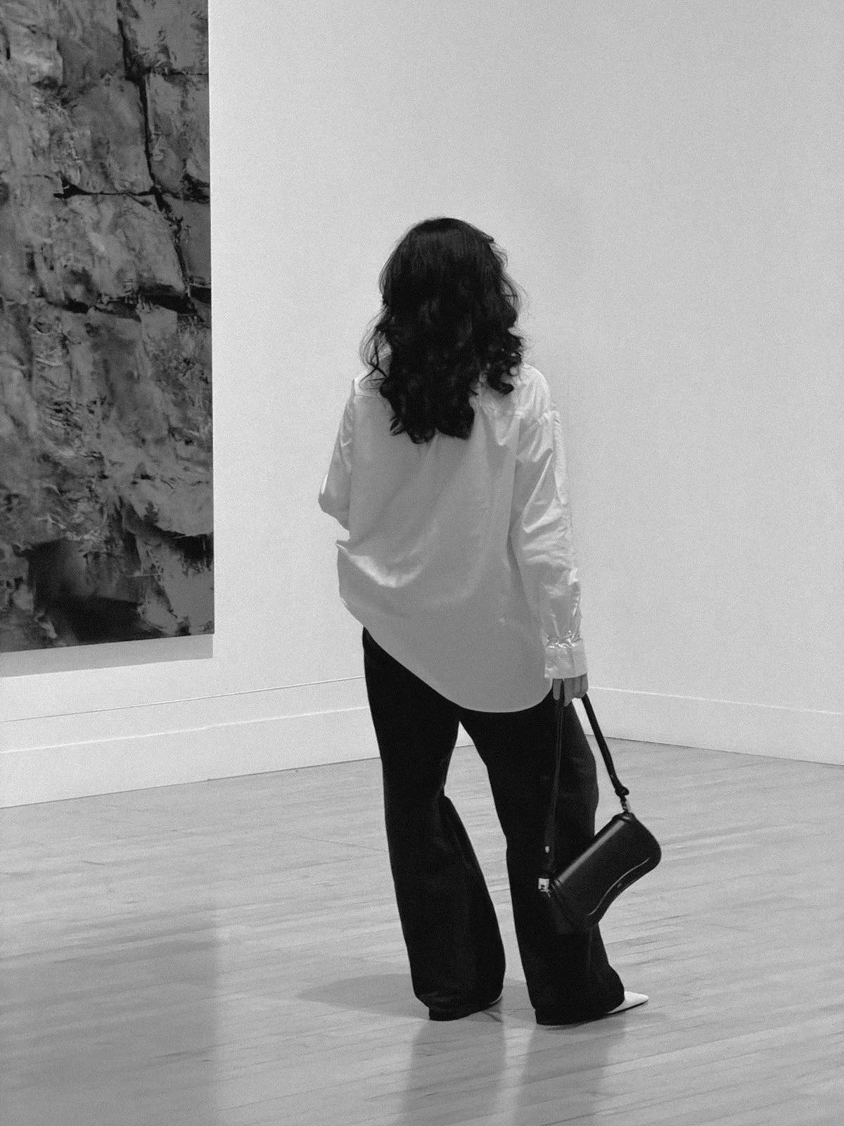 Back view of a woman with curly hair in a white shirt and black pants, holding a black handbag, standing in art gallery with a large painting on the wall.