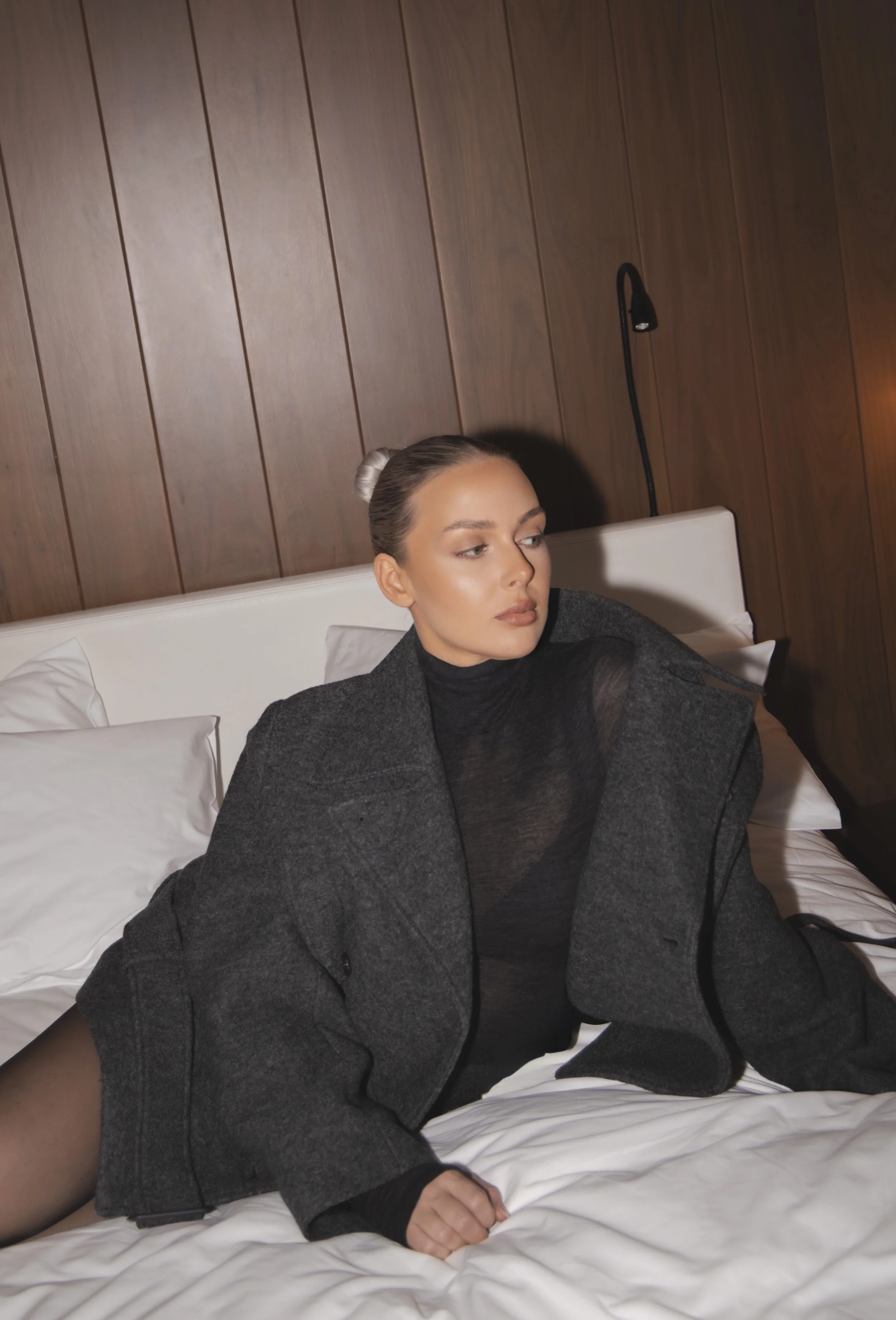 A woman with dark hair styled in a bun, wearing a black turtleneck and a dark gray blazer, sitting on a bed with white sheets and pillows, against a wooden-paneled wall with a black reading lamp.