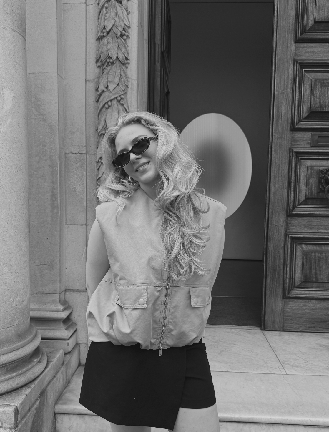 A woman with long wavy hair wearing sunglasses, a light sleeveless vest, and a dark skirt, standing outside a building with ornate columns and a wooden door.