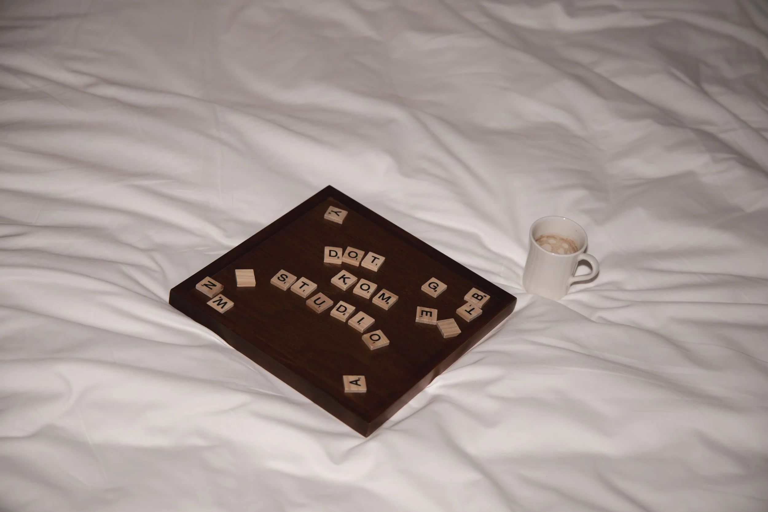 A dark wooden tray on a white bedsheet with scattered Scrabble tiles spelling out words, and a white mug with a hot beverage nearby.