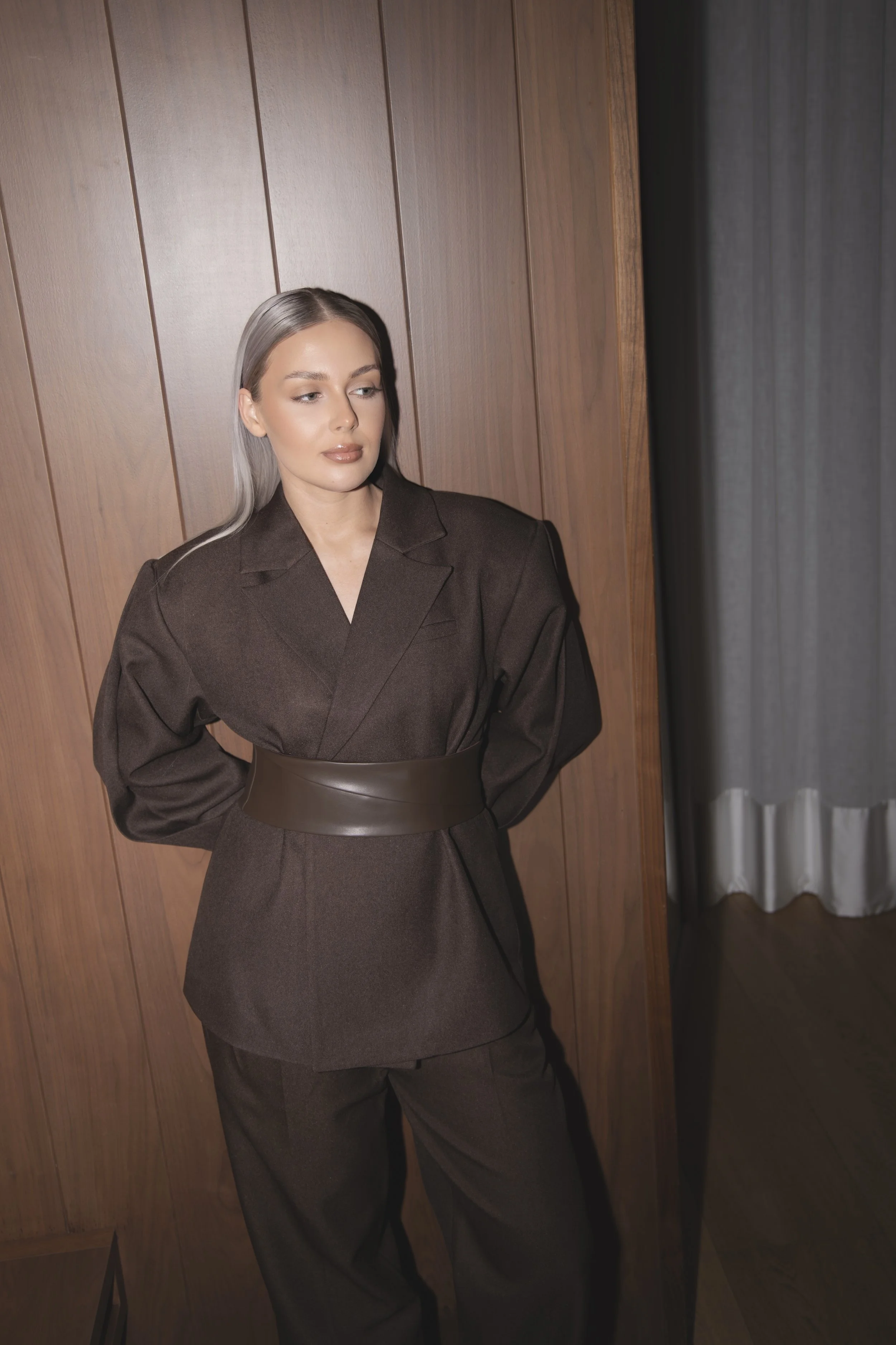A woman with platinum blonde hair in a brown suit with a matching wide belt standing against a wooden-paneled wall.