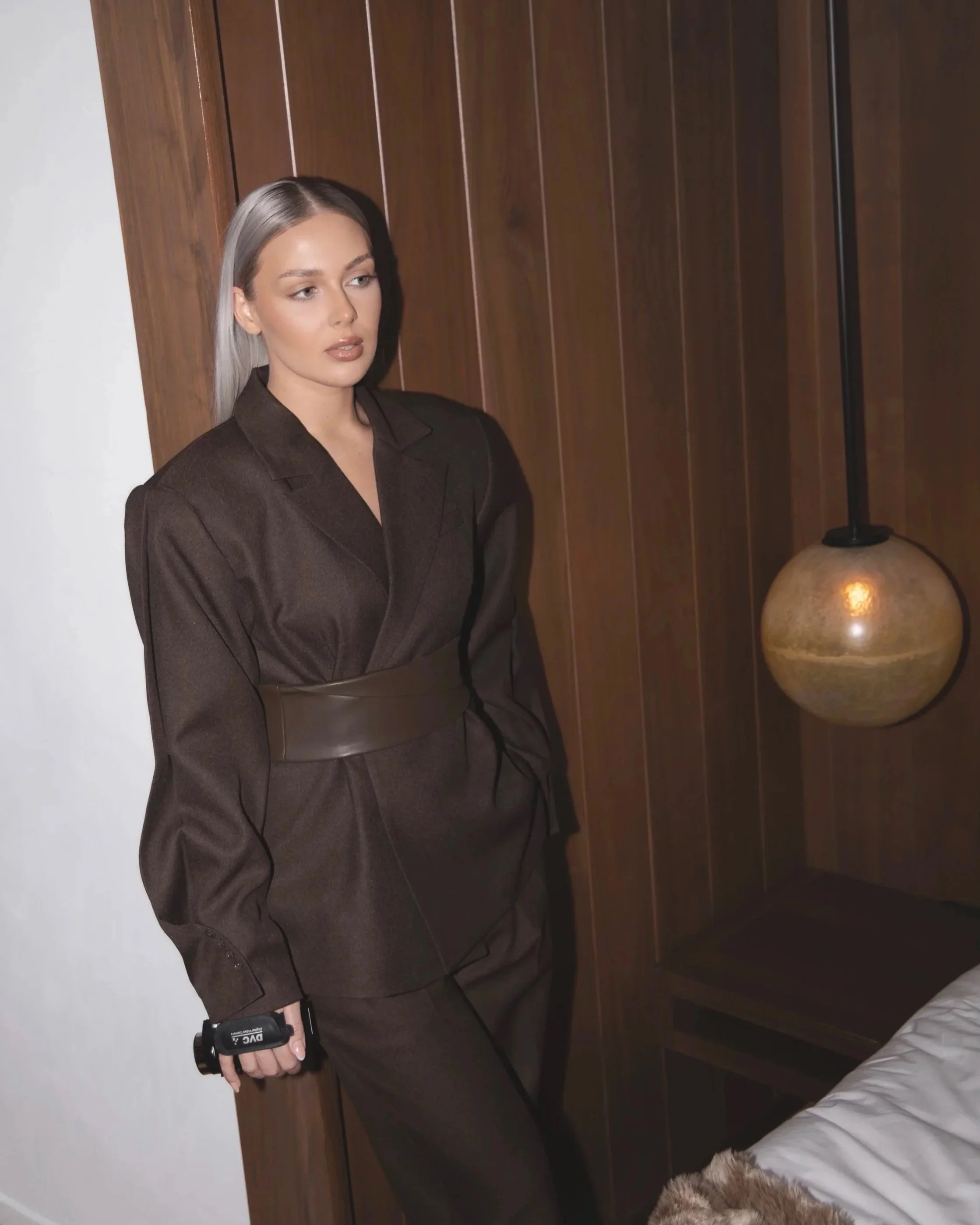 Woman with silver hair wearing a dark brown suit standing beside a wooden wall and a modern wall lamp.