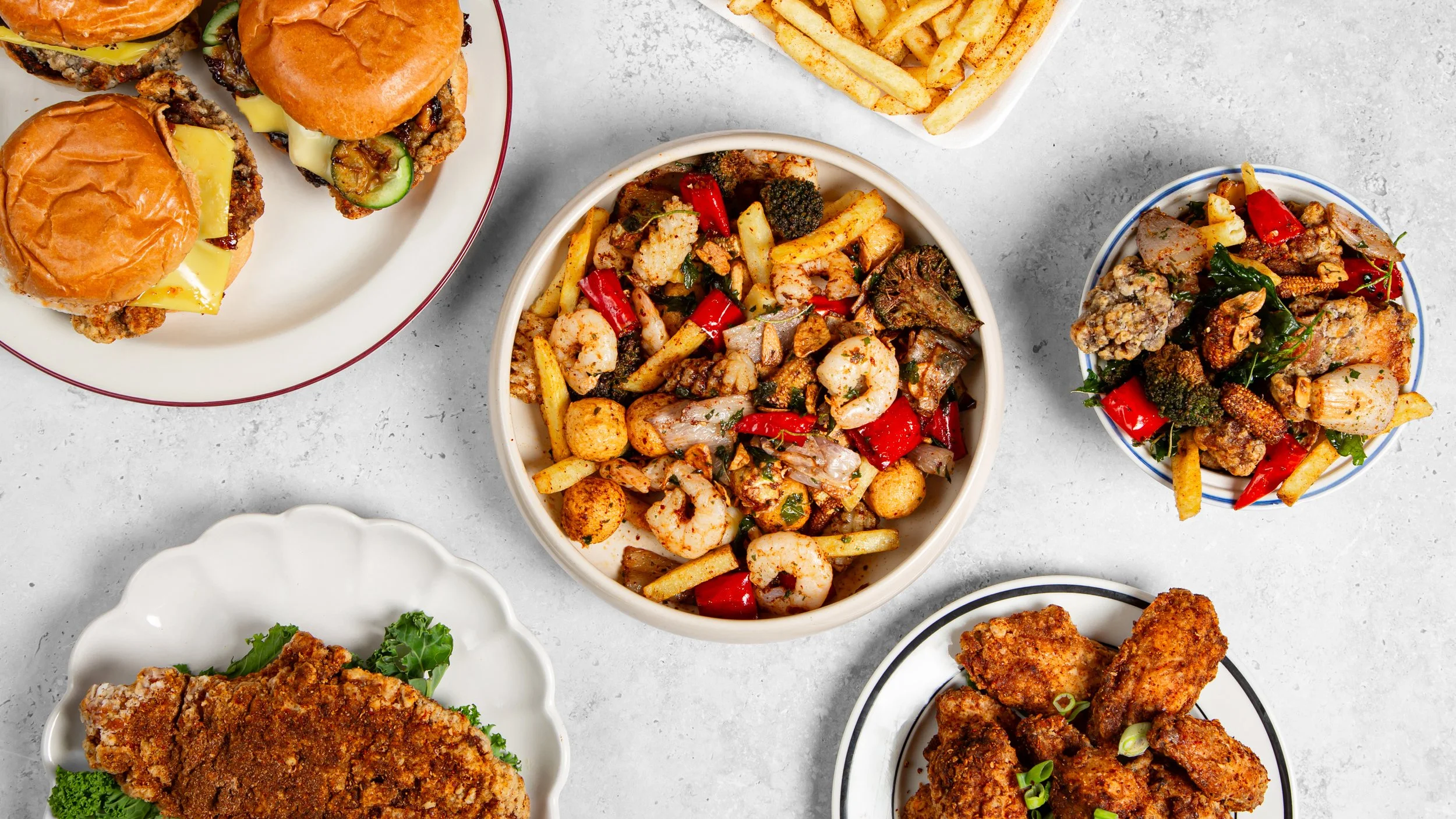 Assorted dishes including fried chicken, prawnsand French fries, fried chicken burgers and Taiwanese Fried Chicken wings on plates and bowls on a gray surface.