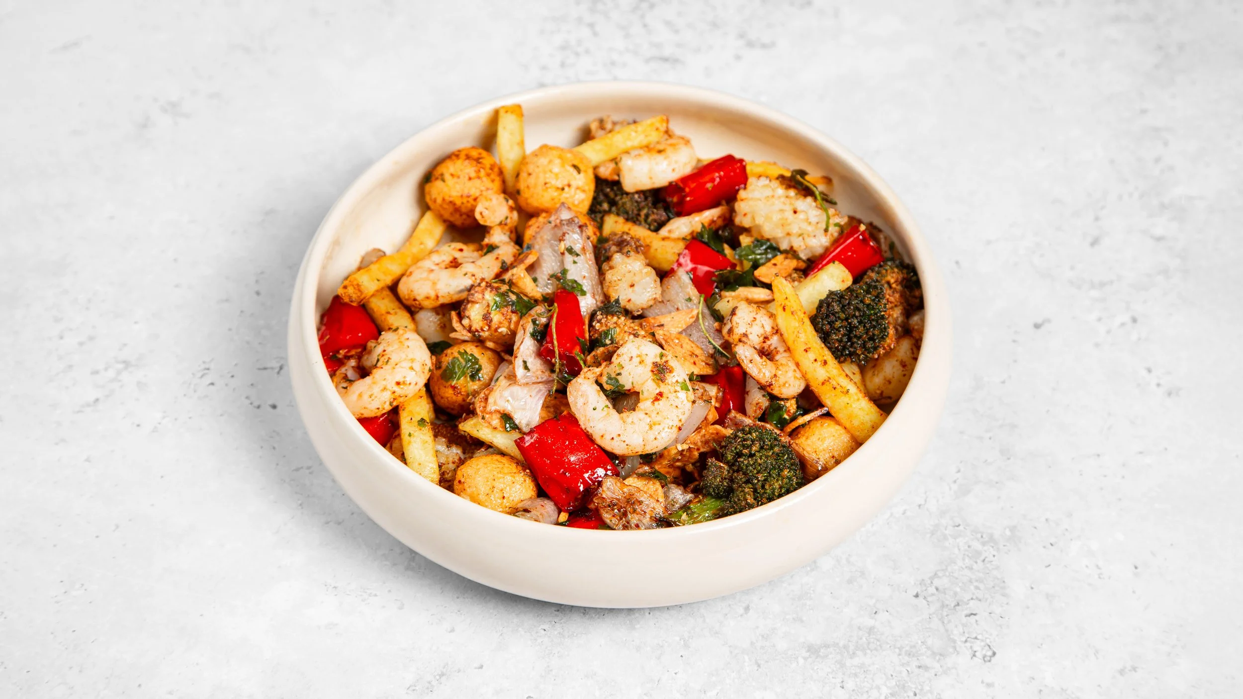 A white bowl filled with a colorful mixture of shrimp, broccoli, red chilli peppers, and French fries also known as Yan Su Ji or an authentic Seafood Spice Bag on a light gray surface.