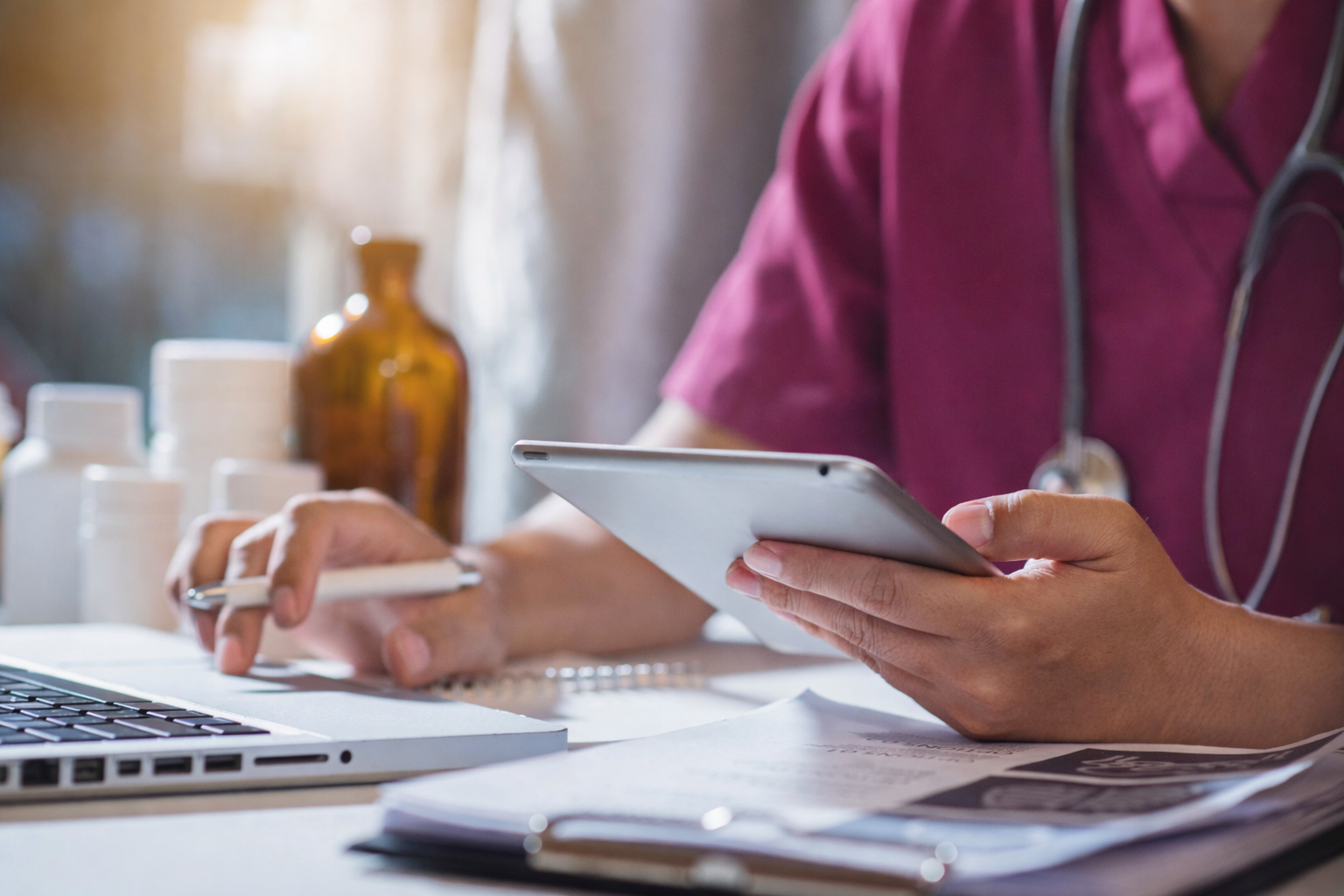 A healthcare professional wearing a stethoscope and burgundy scrubs working at a desk with a laptop, notebook, and medicine bottles.