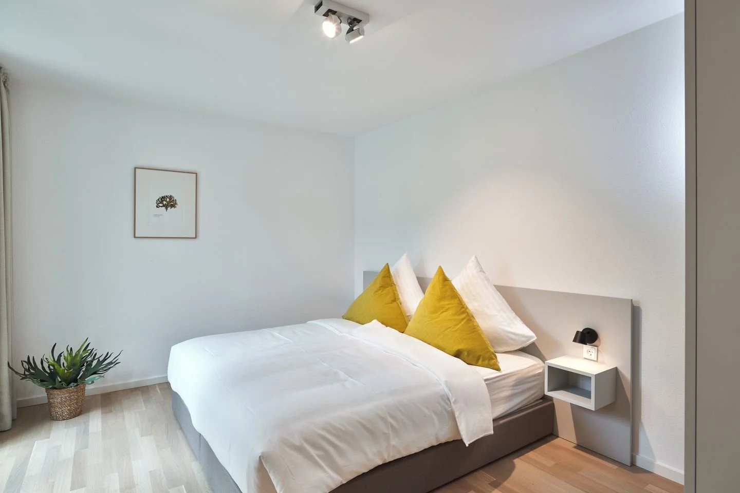 Schlafzimmer mit weißer Wand, Bett mit weißen und gelben Kissen, kleiner Nachttisch mit schwarzen Wandlampe, Pflanze in Korb, Bild an der Wand