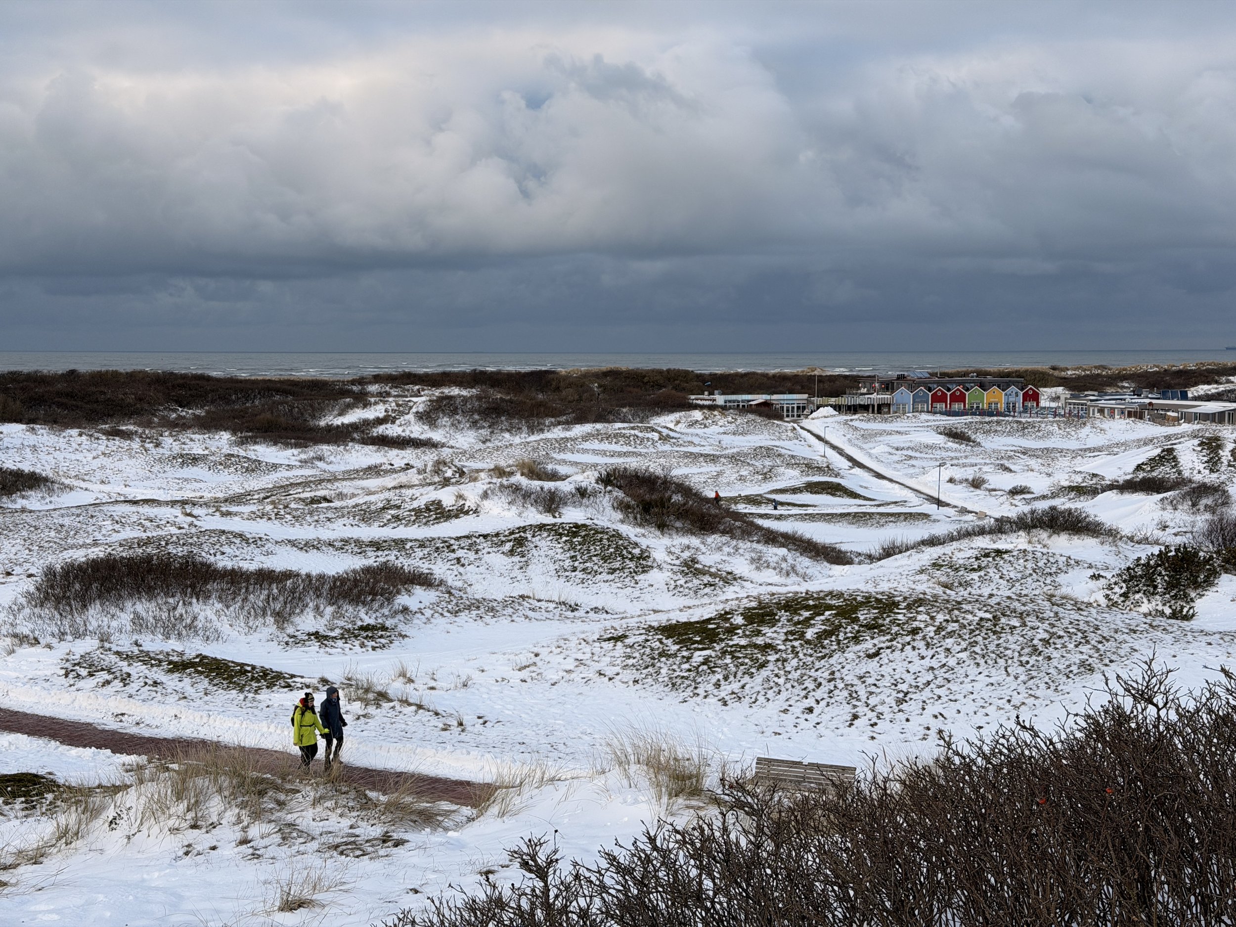 Zwei Personen wandern auf einem schneebedeckten Wanderweg in einer winterlichen Dünenlandschaft mit Häusern im Hintergrund unter einem bewölkten Himmel.