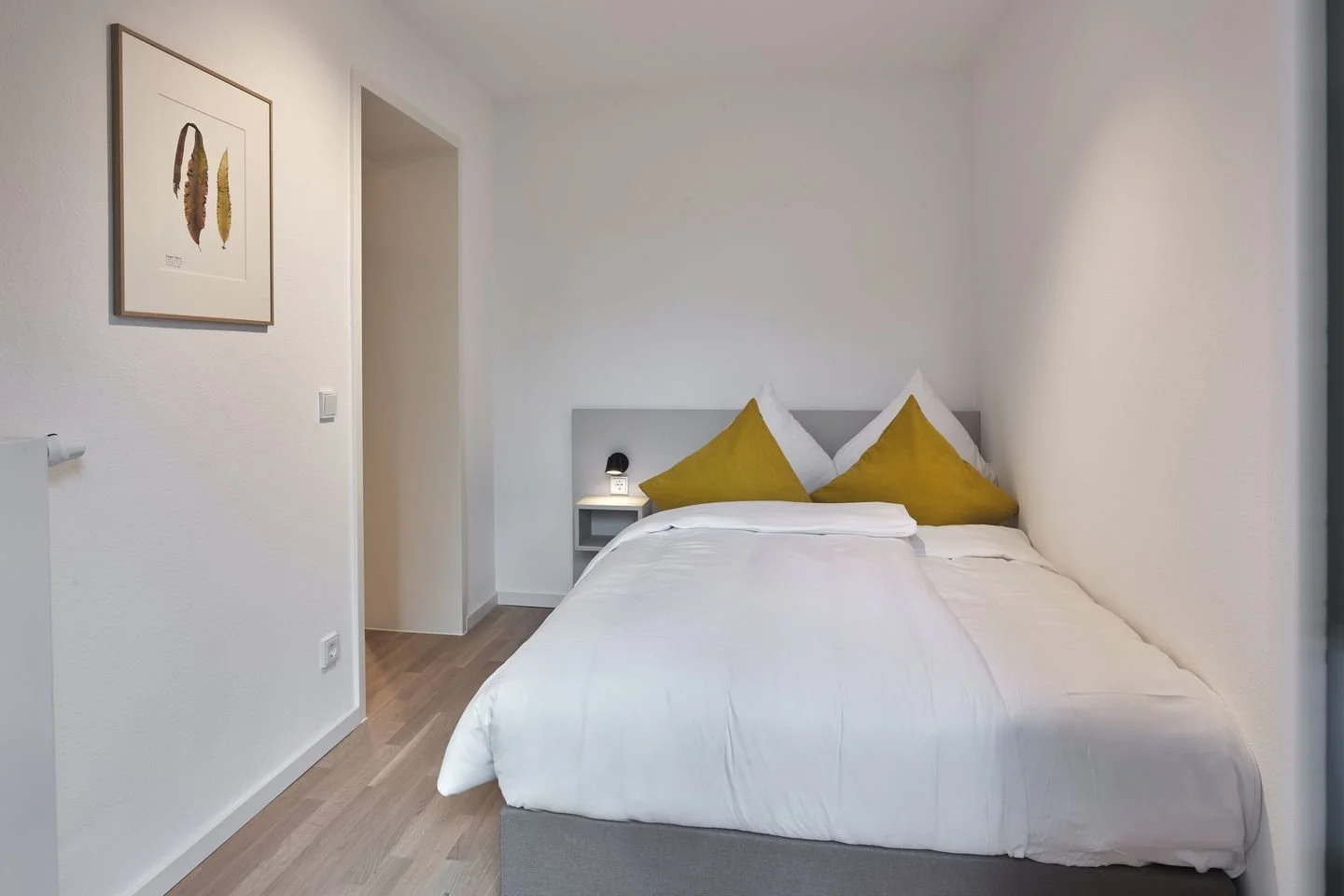 Schlafzimmer mit Doppelbett, weißen Bettwäsche, zwei weißen und zwei senfgelben Kissen, schlichter grauer Kopfteil, Nachttisch mit Nachtischlampe, weißer Wand mit Kunstwerk und Lichtschalter