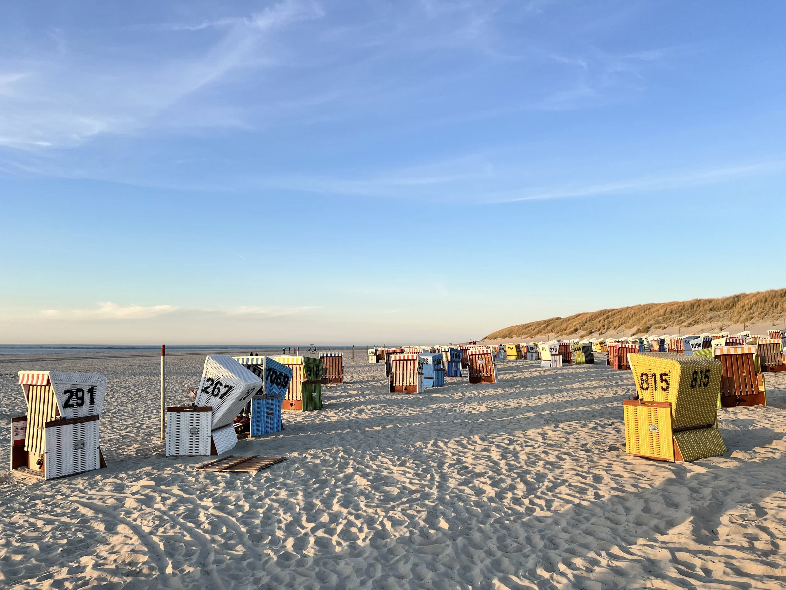 Bunte Strandkörbe auf einem Sandstrand bei Sonnenuntergang mit Dünen im Hintergrund.