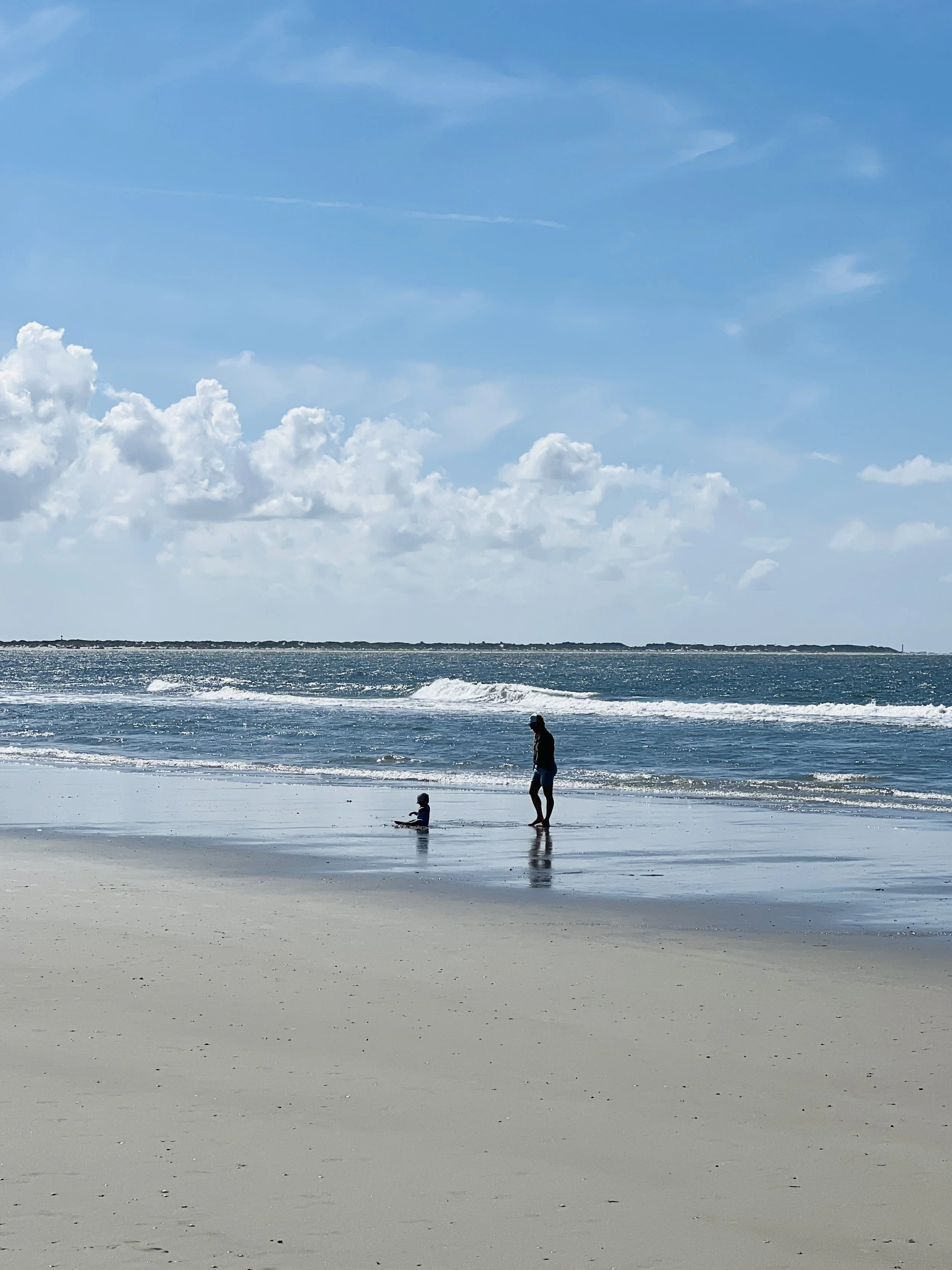 Ein Erwachsener und ein Kind am Strand, im Hintergrund das Meer bei sonnigem Himmel.