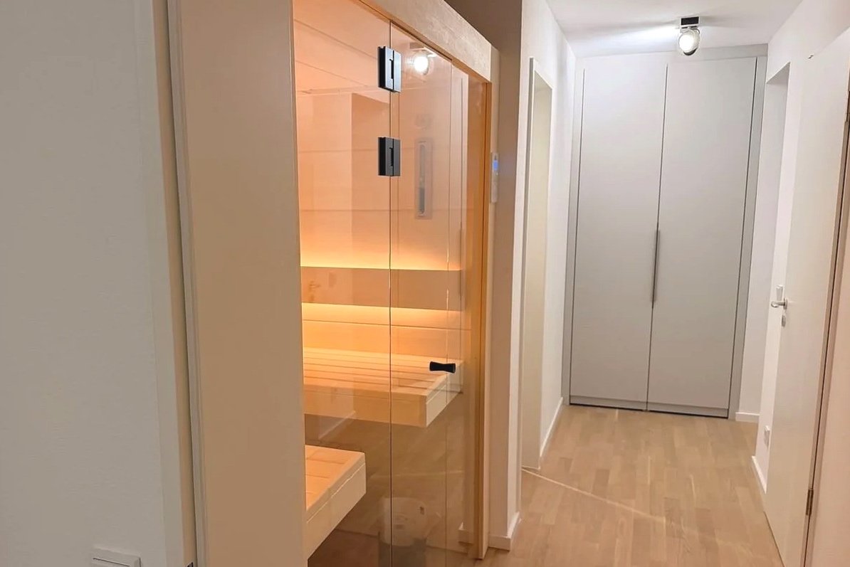 Innenansicht eines Flurs mit einem hölzernen Sauna-Raum mit Glaswand und Jasmin-Leuchte, und einem weißen Schrank. Der Boden ist aus Holz, die Wände sind weiß.